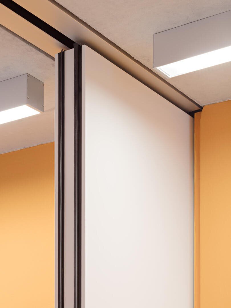 Gros plan sur une cloison coulissante moderne avec un panneau blanc et une moulure noire entre des murs jaunes et orange et sous un plafond en béton avec des plafonniers rectangulaires.
