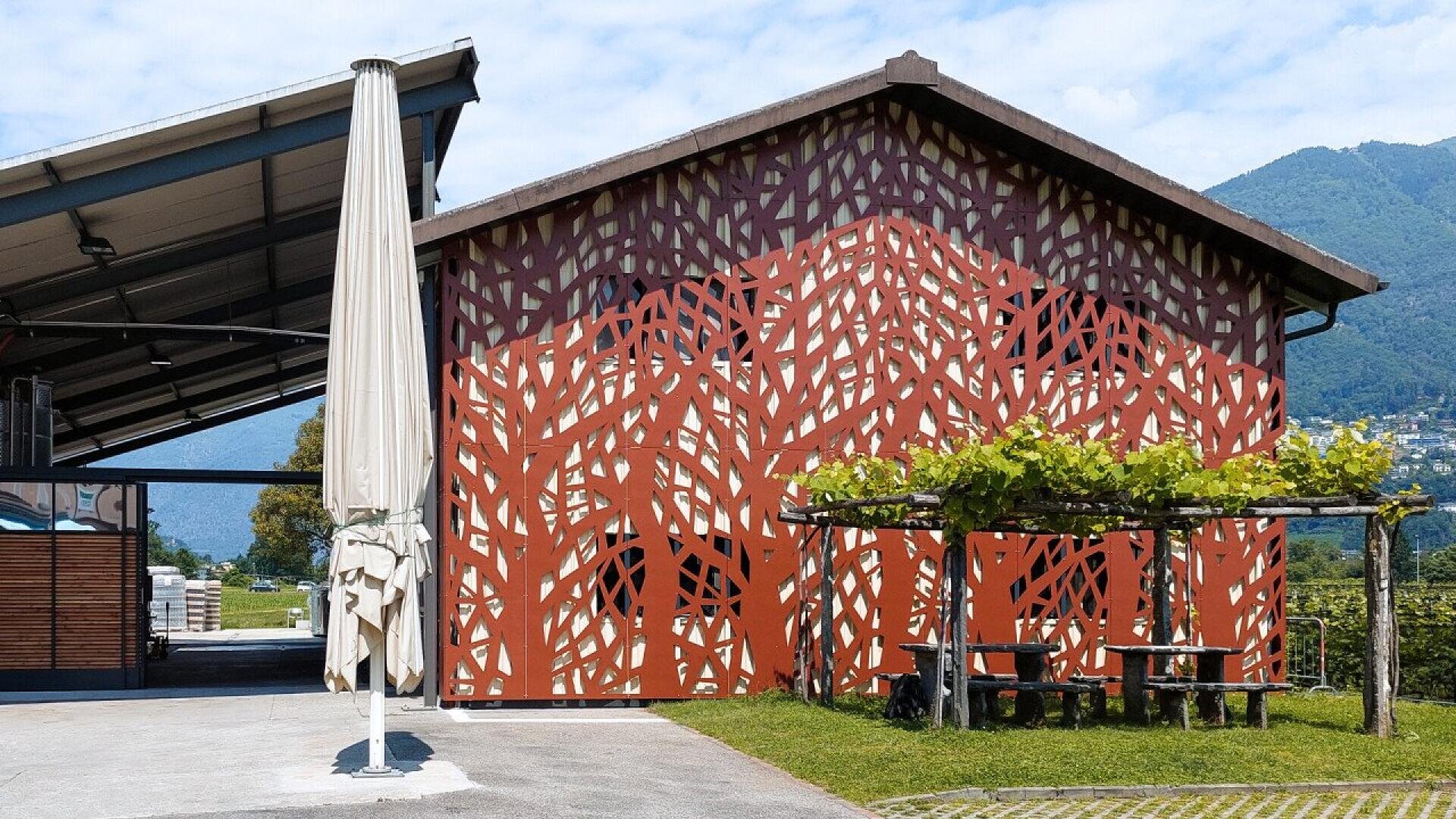 Un petit bâtiment dont la façade est recouverte d'un motif abstrait de treillis rouge se trouve à côté d'un parasol et de tables de pique-nique, avec des montagnes vertes et un ciel bleu en arrière-plan.