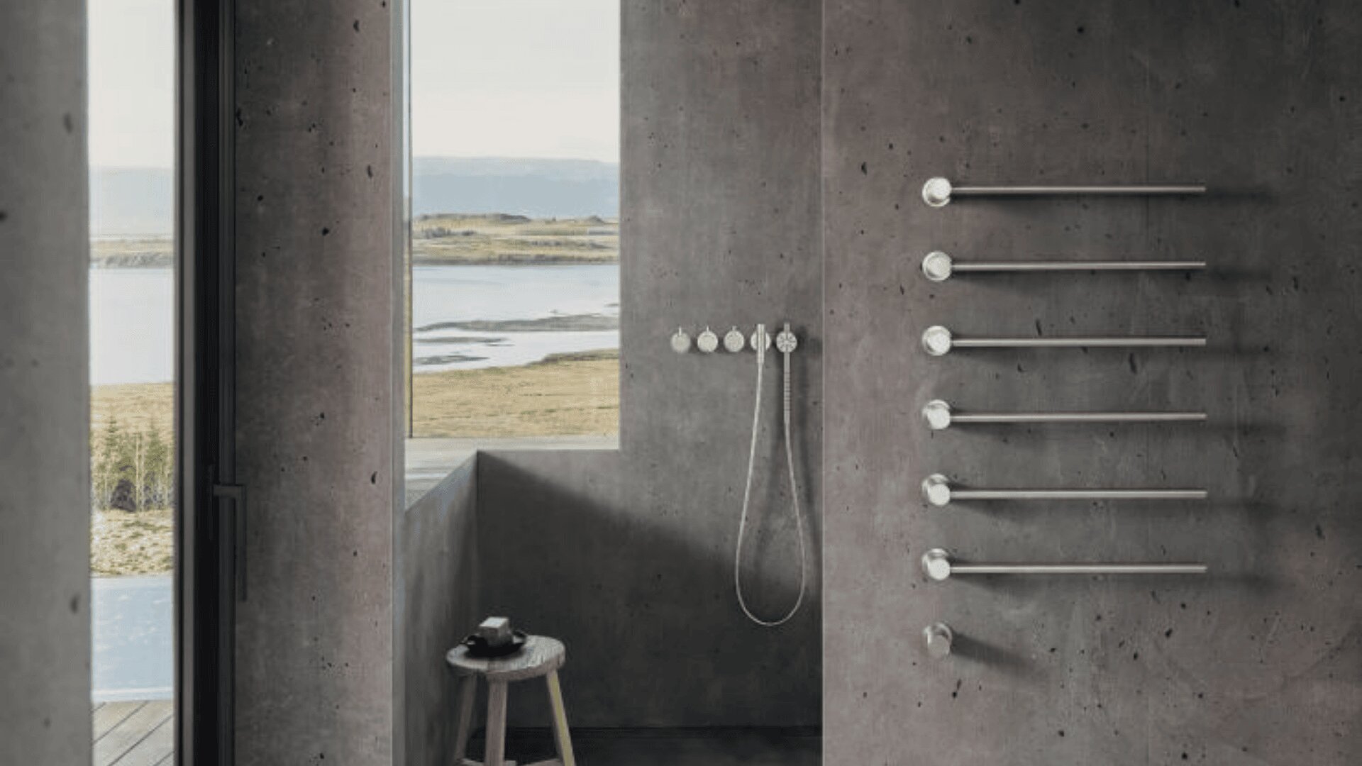 Ein minimalistisches, modernes Badezimmer mit glatten grauen Betonwänden, einer begehbaren Dusche, Wandarmaturen, beheizten Handtuchhaltern, einem kleinen Holzhocker und einem großen Fenster mit Blick auf eine ruhige Naturlandschaft mit Wasser und Hügeln.