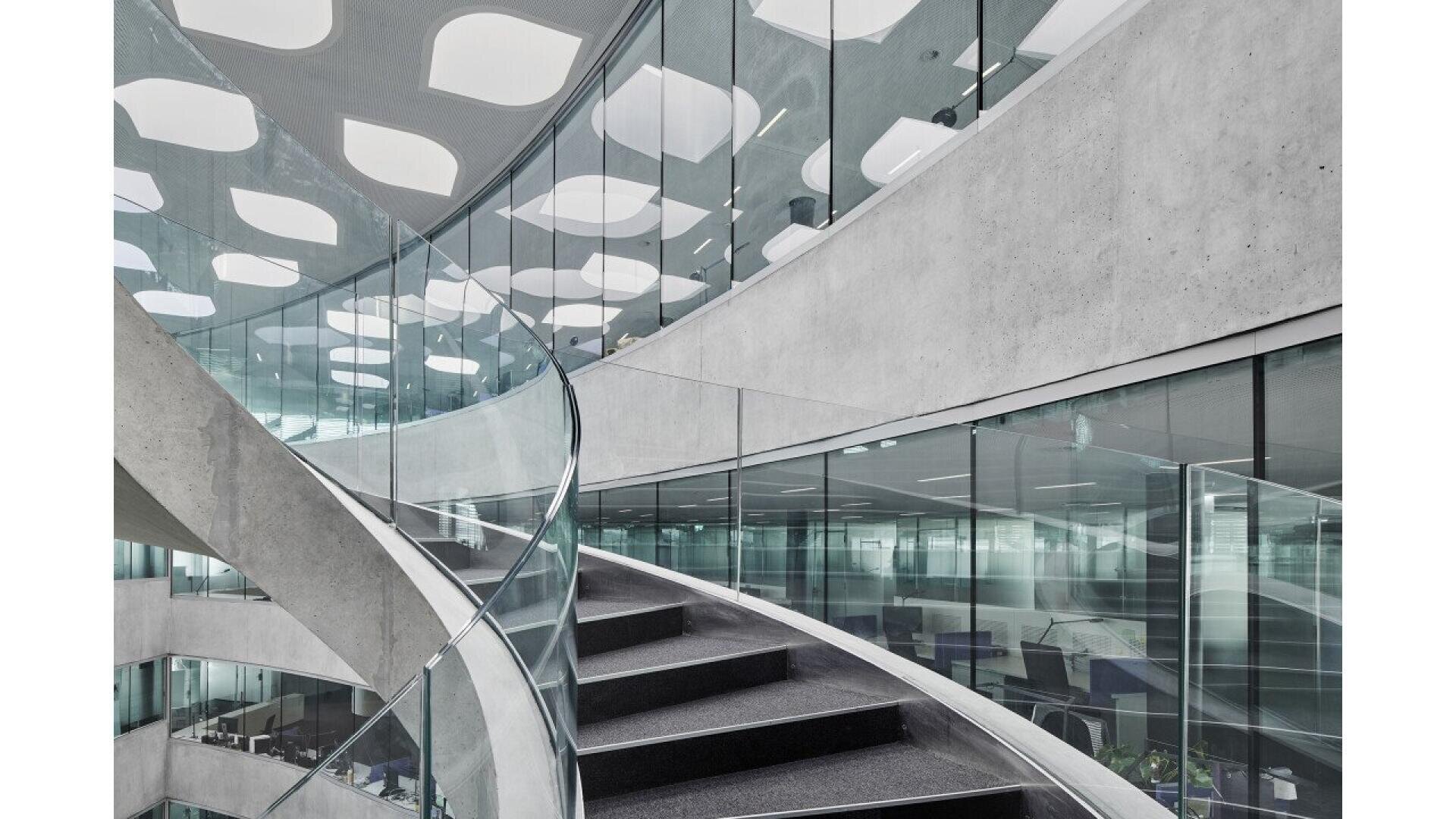 Un intérieur de bureau moderne avec un escalier incurvé et vitré, des murs en béton et de grandes fenêtres. Le plafond comporte des panneaux lumineux géométriques et les bureaux sont visibles à travers le verre à l'étage inférieur.