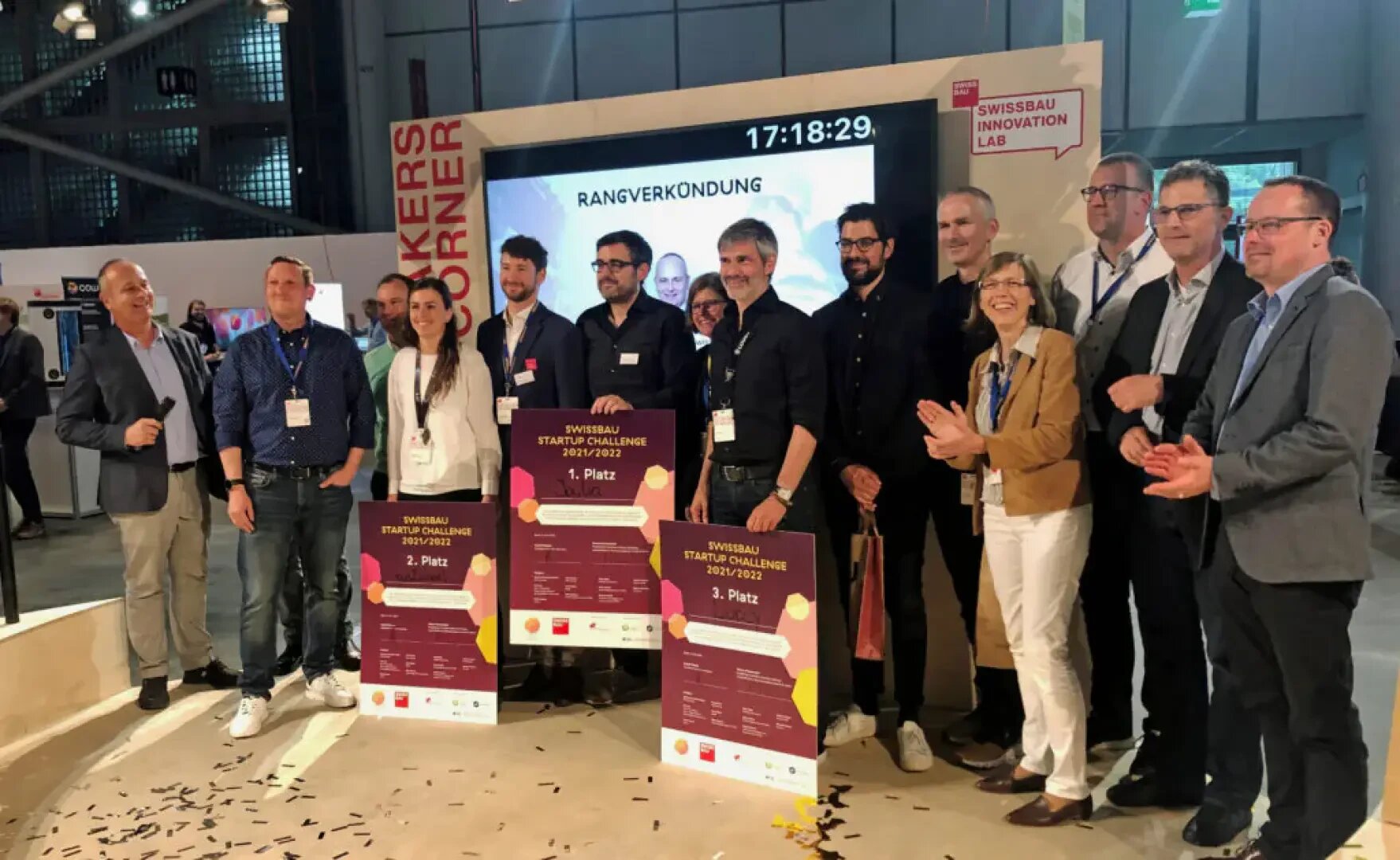 Eine Gruppe von Personen steht in einem Innenraum, einige halten grosse Preise und Urkunden in der Hand. Sie posieren und lächeln für ein Foto an der Swissbau Startup Challenge Veranstaltung, mit einem Bildschirm und Bannern im Hintergrund.