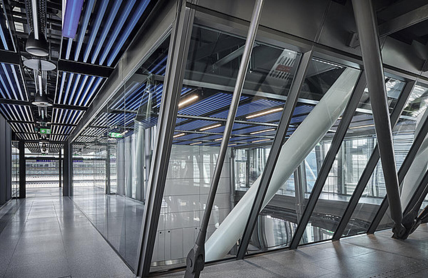 Modernes Interieur eines Gebäudes mit Glaswänden, schrägen Stahlträgern und einer Decke mit blau-schwarz gestreiften Paneelen. Helles natürliches Licht fällt durch große Fenster ein und verleiht dem Raum einen offenen, industriellen Charakter.