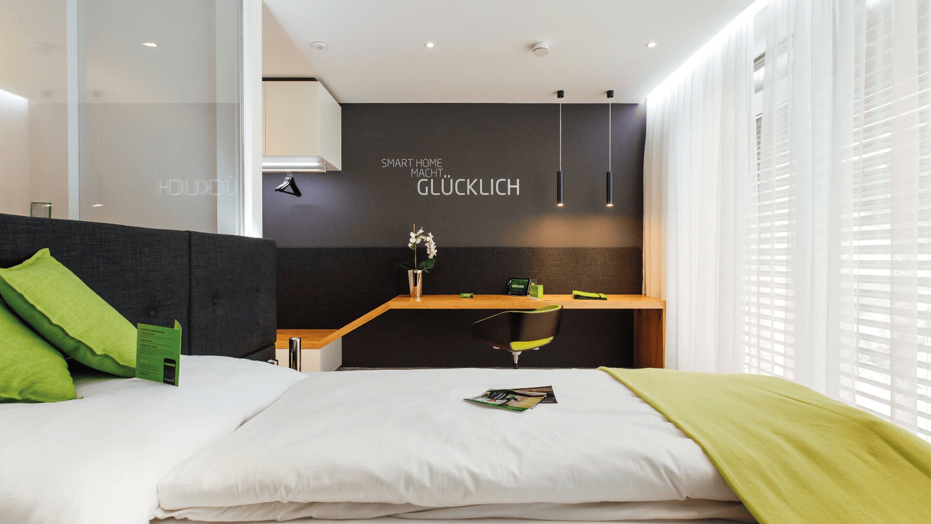 Modernes Hotelzimmer mit einem ordentlich gemachten Bett, grünen Akzenten auf Kissen und Decke, einem Holzschreibtisch mit Stuhl und einer dunklen Akzentwand mit der Aufschrift "SMART HOME MACHT GLÜCKLICH". Licht strömt durch durchsichtige Vorhänge herein.