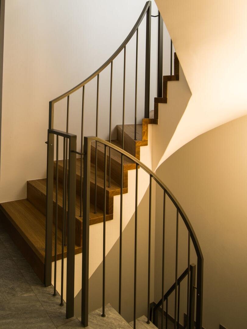 Sonnenlicht beleuchtet eine moderne Holztreppe mit schwarzen Metallgeländern, wirft markante Schatten auf die weißen Wände und unterstreicht das geschwungene Design der Treppe.