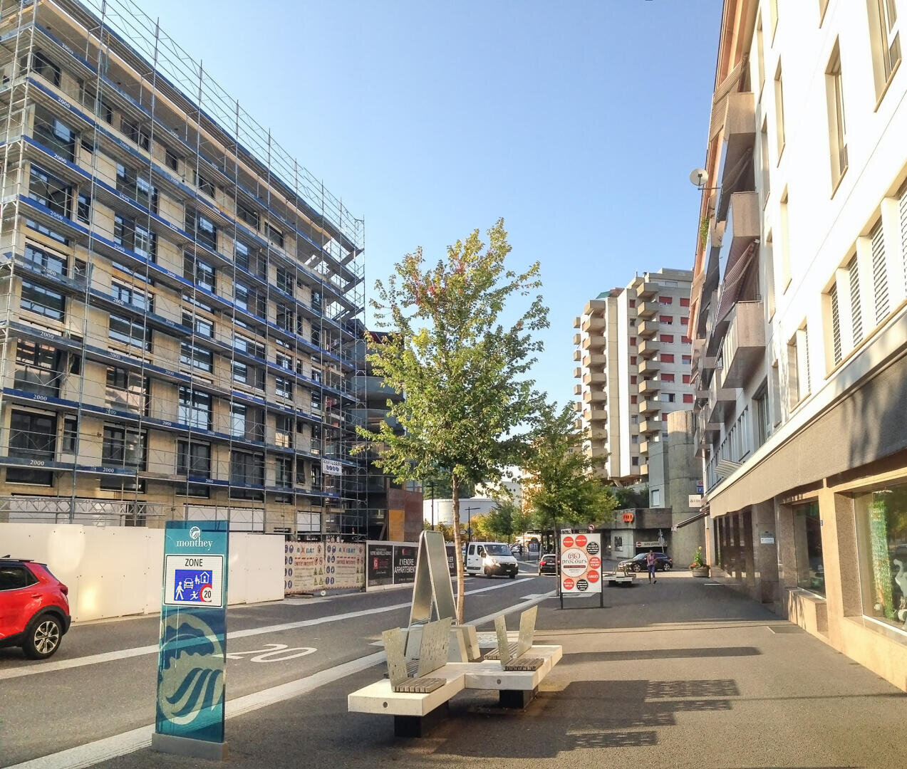 Eine sonnige Stadtstraße mit modernen Gebäuden, eines davon im Bau, gesäumt von Gerüsten. Auf dem Gehweg sind ein Baum, Bänke, geparkte Autos und Verkehrsschilder zu sehen. Schatten und klarer Himmel lassen auf Morgen oder Nachmittag schließen.