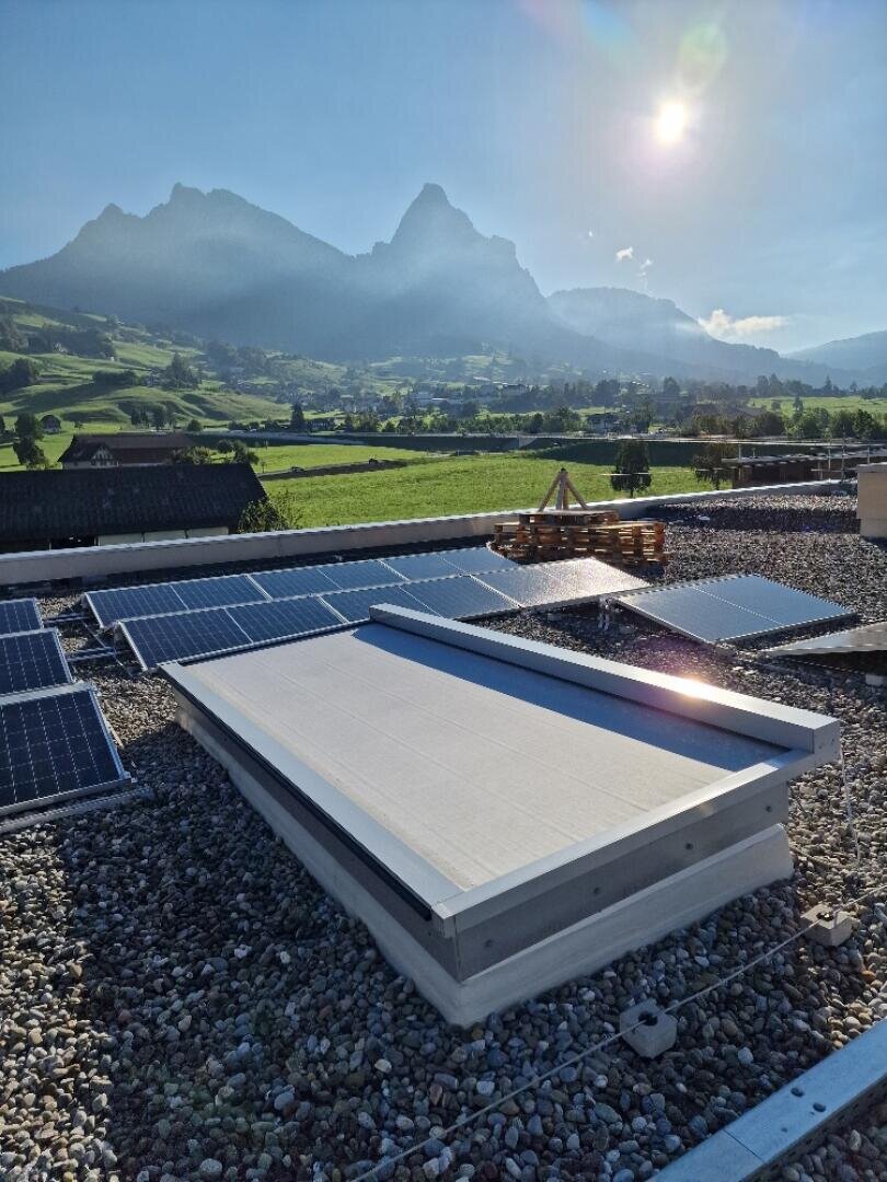 Sonnenkollektoren auf einem Kiesdach mit Bergen und einem sonnigen Himmel im Hintergrund, grünen Feldern und einem kleinen Dorf in der Ferne.