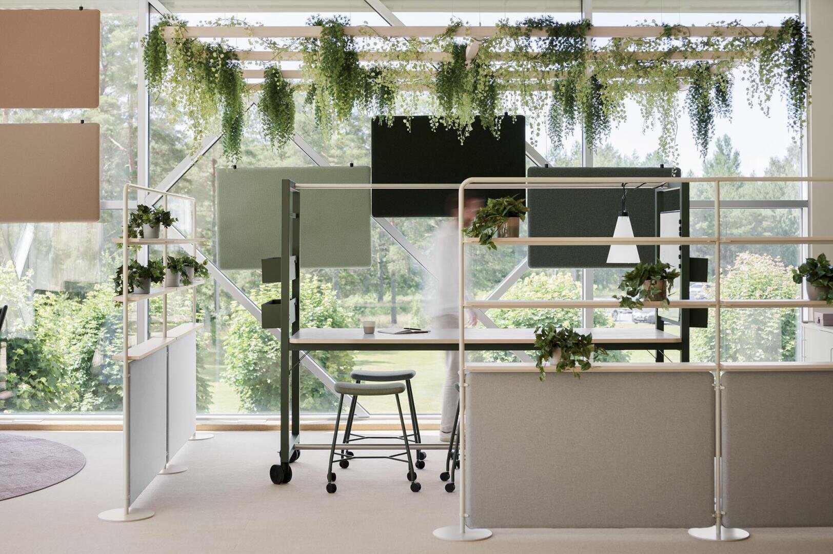 Un espace de travail de bureau moderne avec des bureaux individuels séparés par des cloisons et décorés de plantes vertes suspendues et en pot. De grandes fenêtres donnent sur la verdure luxuriante à l'extérieur et créent une atmosphère lumineuse et aérée.