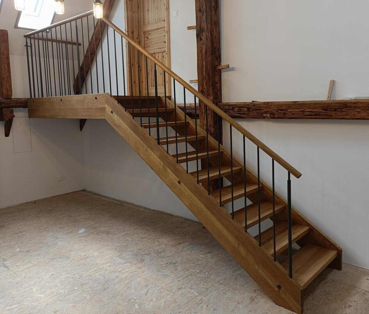 Eine Holztreppe mit Metallgeländer führt zu einem oberen Stockwerk in einem Raum mit unbearbeitetem Fußboden und freiliegenden Holzbalken an den Wänden. Der Raum scheint gerade renoviert zu werden.