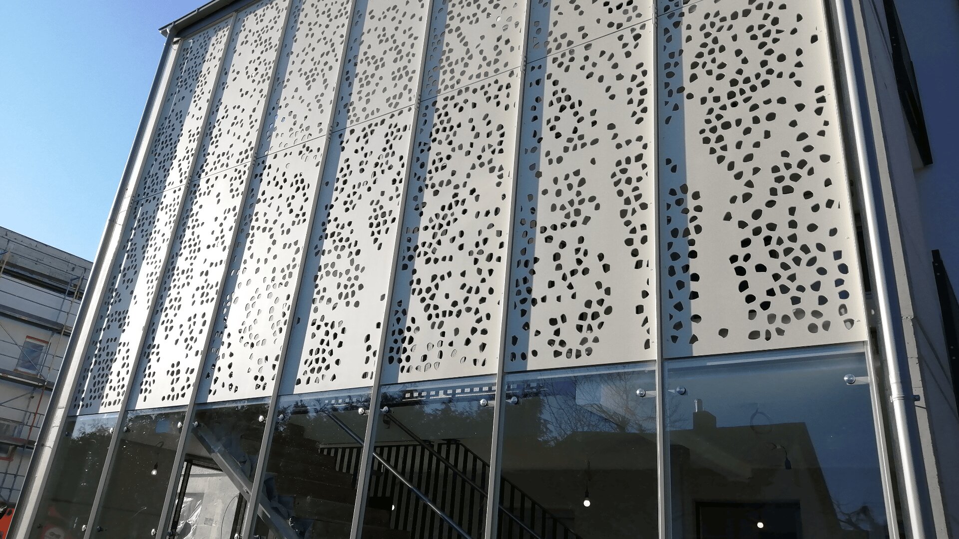 Eine moderne Gebäudefassade mit großen Glasscheiben im unteren Bereich und oberen Scheiben mit einem abstrakten Muster aus unregelmäßig geformten Ausschnitten. Treppen und Spiegelungen sind durch das Glas sichtbar.