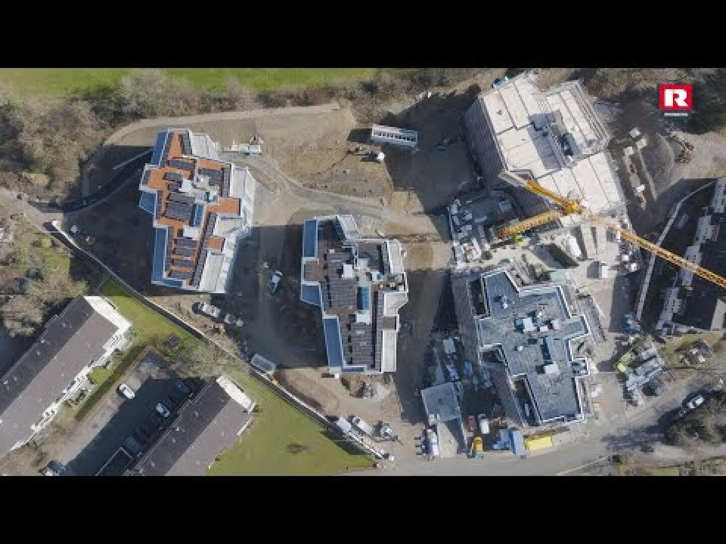 Luftaufnahme einer Baustelle mit mehreren Gebäuden in verschiedenen Stadien der Fertigstellung, umgeben von Erde, Maschinen und Bäumen. Auf der rechten Seite des Bildes ist ein gelber Kran zu sehen.