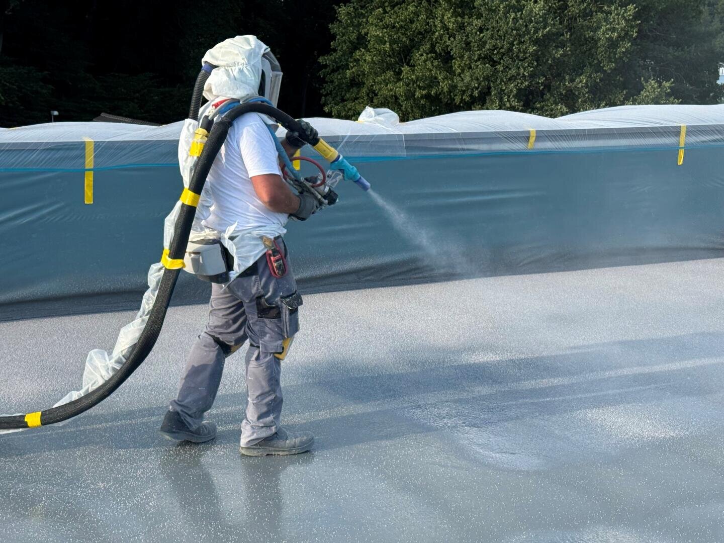 Une personne en tenue de protection pulvérise un revêtement sur une grande surface plane, peut-être un toit, à l'aide d'un tuyau relié à un réservoir de sac à dos. Des bâches en plastique et des arbres sont visibles en arrière-plan.