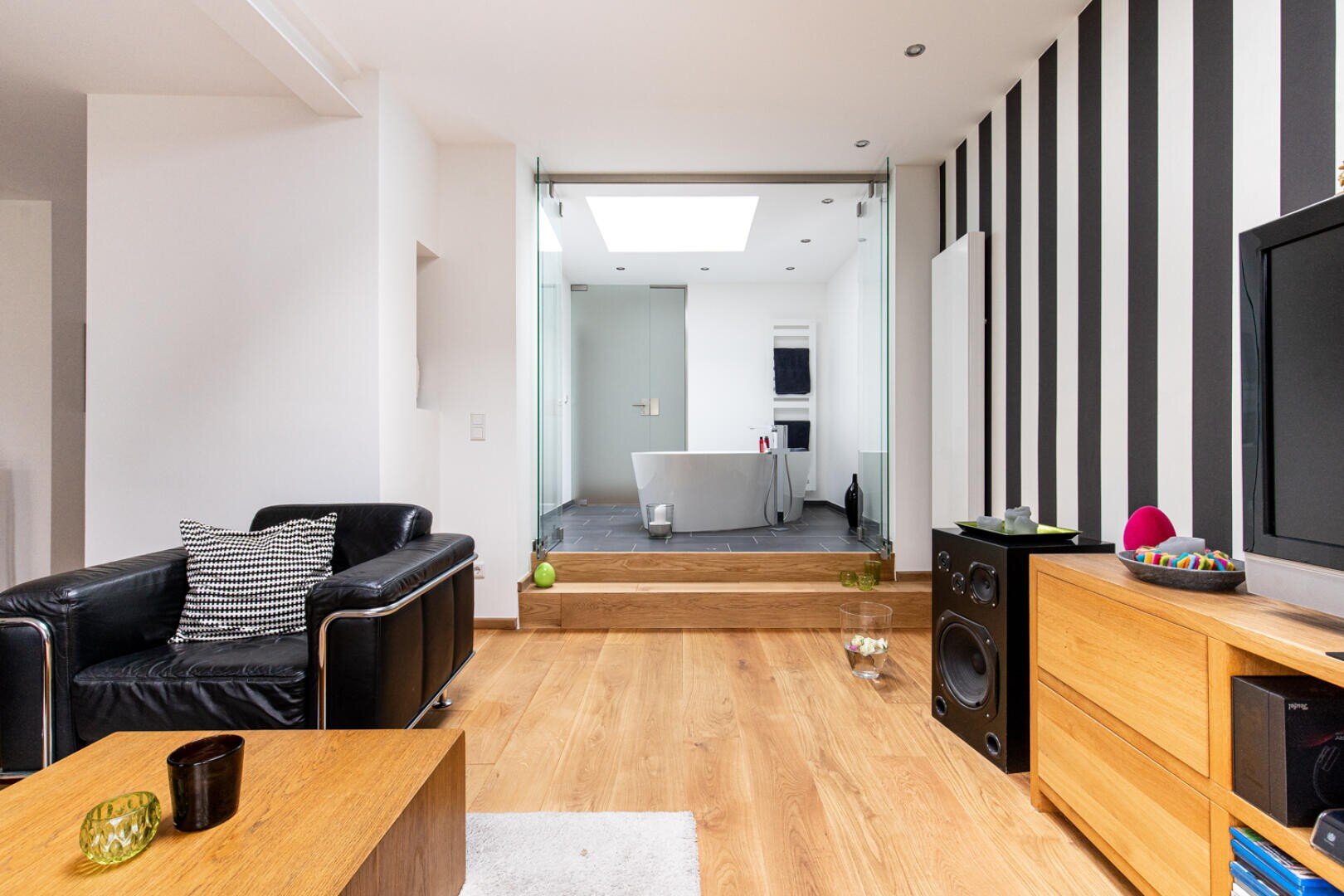 Modernes Wohnzimmer mit Holzfußboden, schwarzem Ledersessel, Holzmöbeln und einem Fernseher. Schwarz-weiß gestreifte Akzentwand auf der rechten Seite. Auf der Rückseite ist ein kleiner erhöhter Bereich mit einem Schreibtisch und Glastüren zu sehen.
