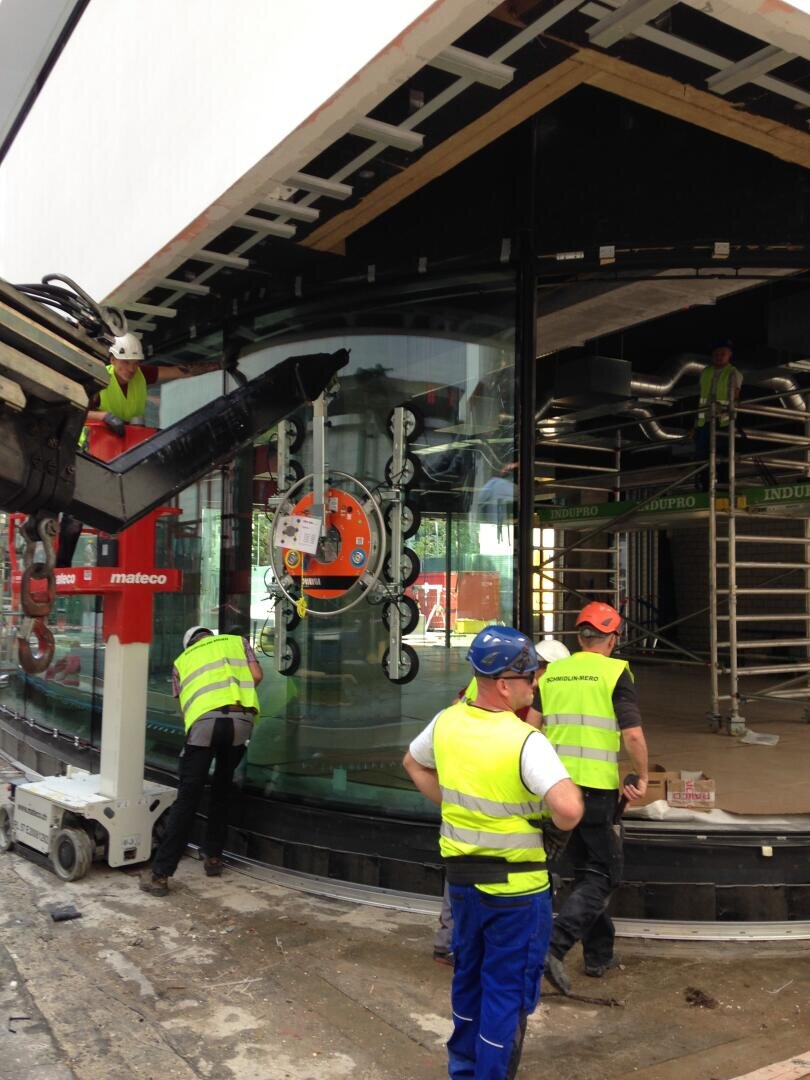 Bauarbeiter montieren auf einer Baustelle mit Hilfe von Hebezeugen eine große gebogene Glasscheibe. Arbeiter mit Sicherheitswesten und Helmen führen das Glas, während andere im Inneren auf Gerüsten arbeiten.
