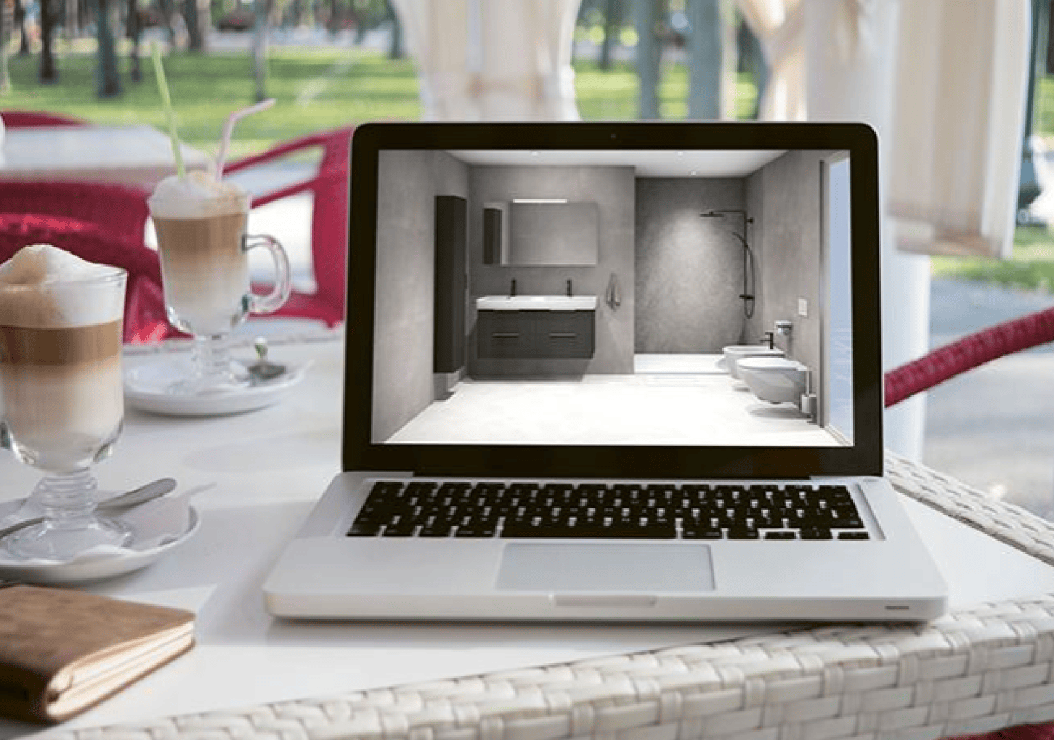 Ein Laptop auf einem weißen Tisch zeigt auf seinem Bildschirm ein modernes Badezimmerdesign. Neben dem Laptop stehen zwei Milchkaffees mit Strohhalmen, eine Brieftasche und eine Brille. Die Szene scheint in einem hellen Café im Freien zu spielen.