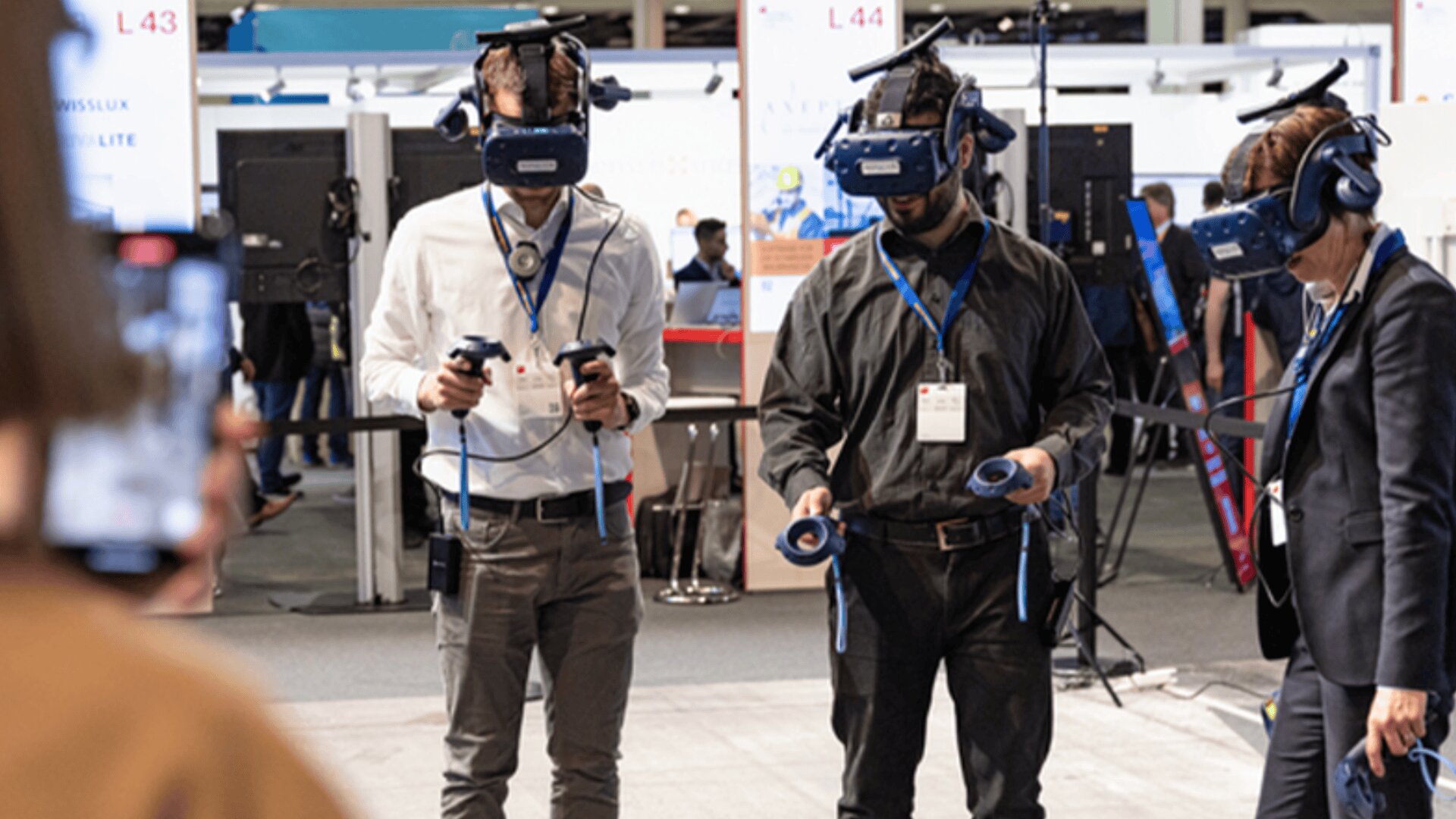 Drei Personen, die VR-Headsets tragen und Controller in der Hand halten, interagieren mit einem virtuellen Erlebnis bei einer Indoor-Veranstaltung. Eine Person im Vordergrund macht ein Foto oder nimmt sie mit einem Smartphone auf.