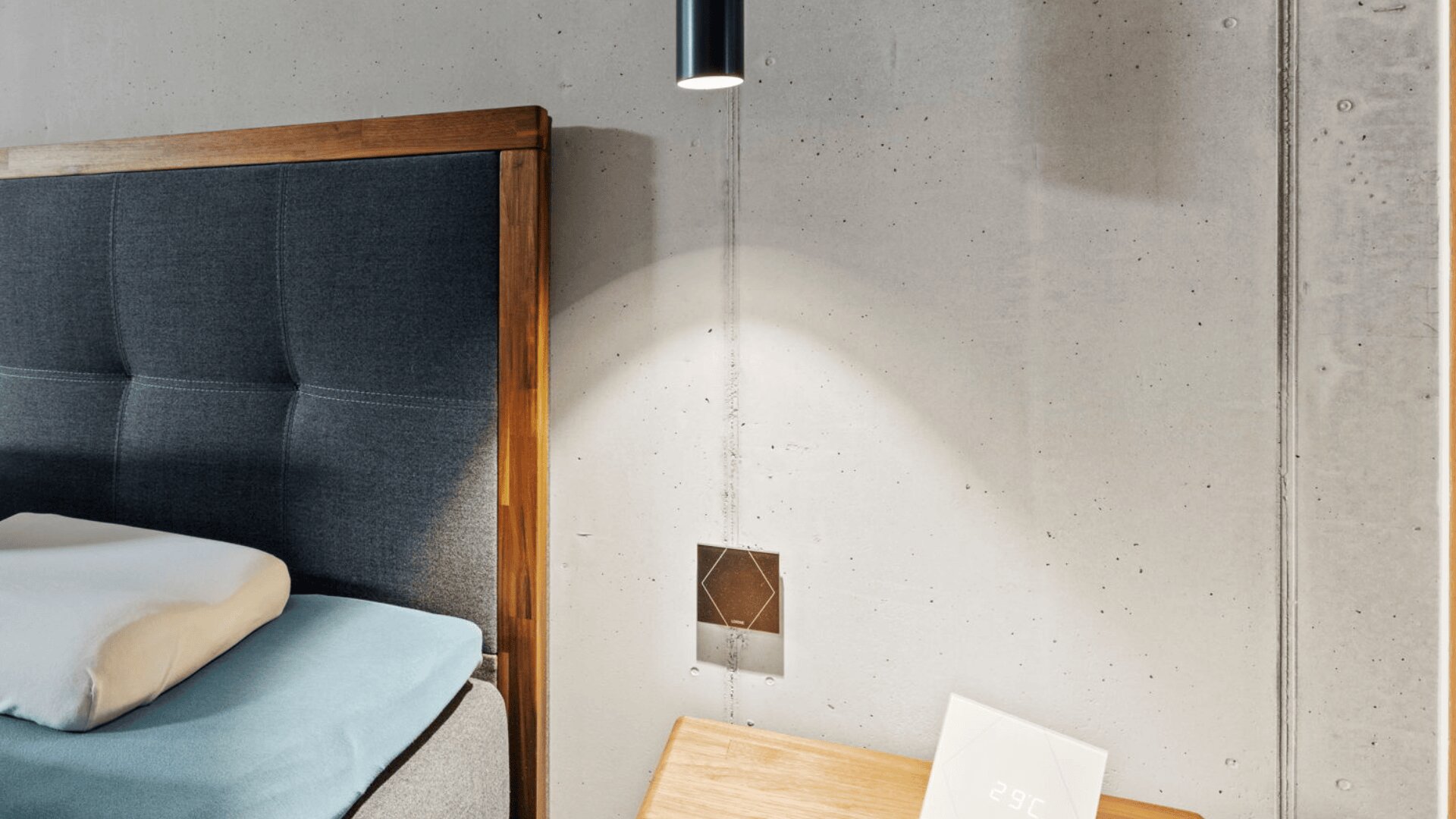 Ein modernes Schlafzimmer mit einem holzgerahmten, dunkelgrauen Kopfteil, einem ordentlich gemachten Bett mit hellblauer Decke, einem Nachttisch, einer Digitaluhr, die 25°C anzeigt, und einer Betonwand mit einer Hängelampe darüber.