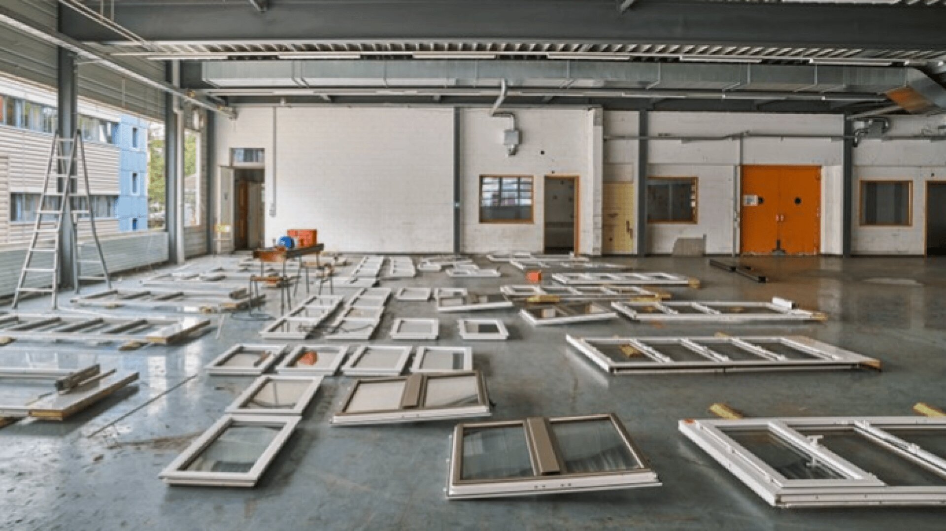 Großer Industrieraum mit vielen Fensterrahmen, die auf dem Boden liegen, einige mit Glasscheiben. An der hinteren Wand befinden sich Fenster und Türen, links eine Leiter und ein paar Werkzeuge und Vorräte.