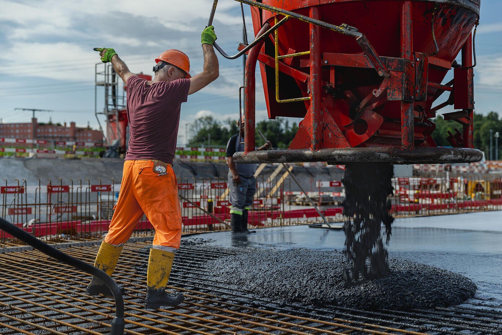 Un ouvrier du bâtiment, vêtu d'un pantalon orange et d'un casque de protection, pilote une grande machine à couler le béton rouge, avec laquelle il dirige le béton mouillé sur un échafaudage d'armature sur un chantier. Un autre ouvrier est visible à l'arrière-plan.
