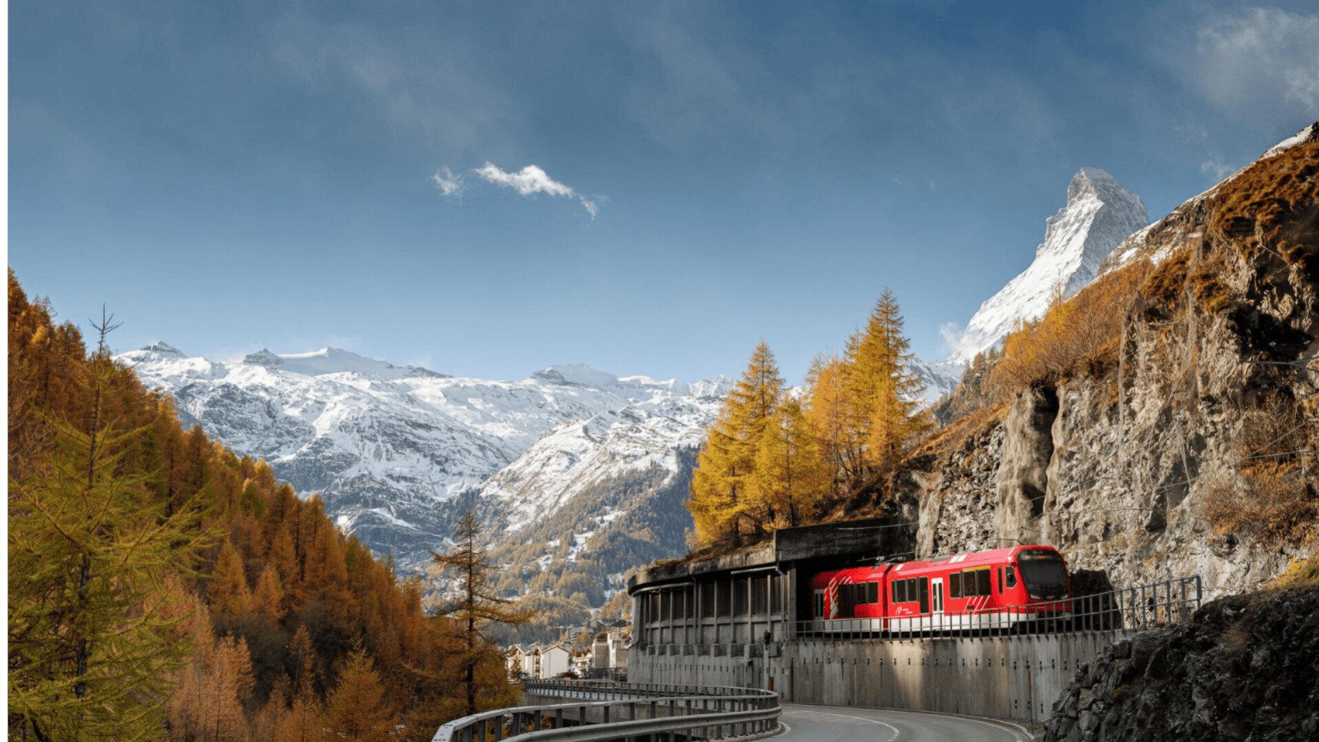Ein roter Zug fährt auf einer geschwungenen Bergstrecke, die von herbstlichen Bäumen gesäumt ist. Im Hintergrund erheben sich schneebedeckte Gipfel, darunter das Matterhorn, unter einem klaren blauen Himmel.