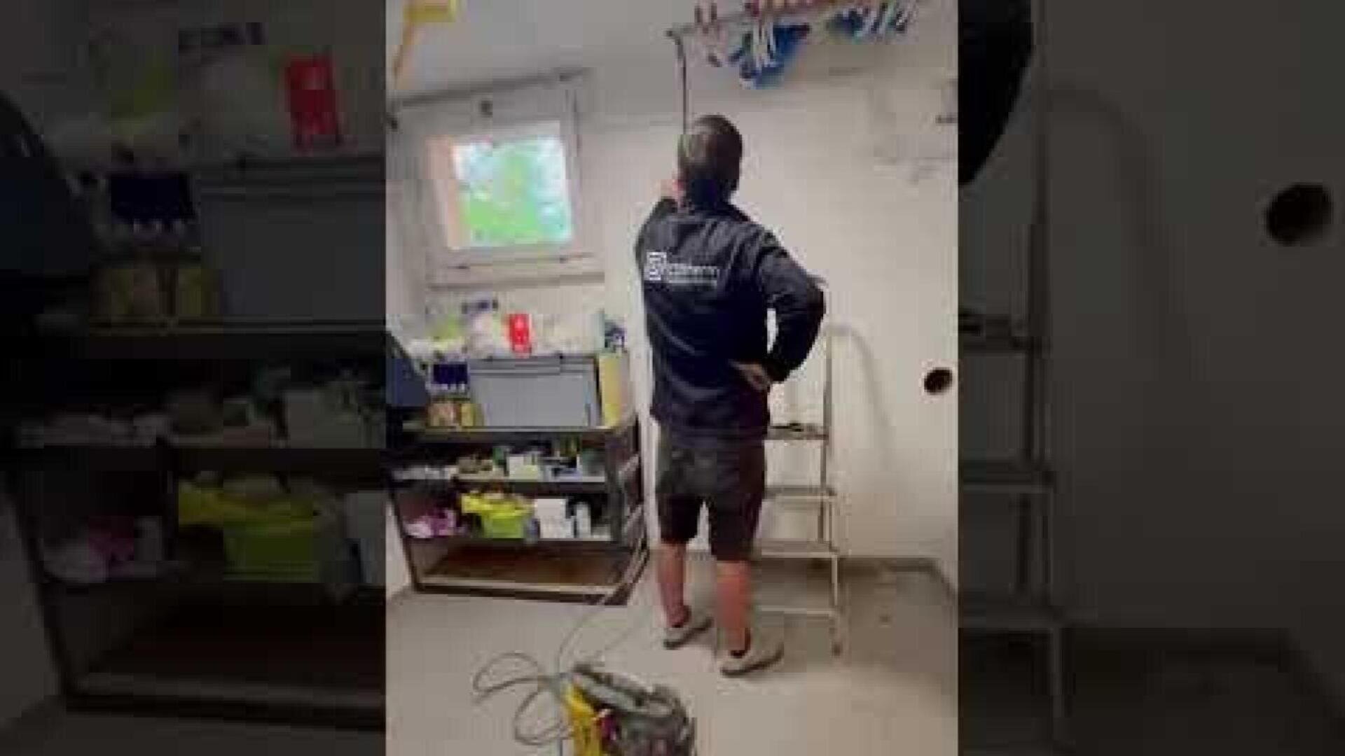 Une personne en tenue de travail se tient sur le sol à côté d'un petit escabeau dans une buanderie et utilise un outil. En arrière-plan, on voit des étagères avec différents matériaux et une fenêtre.
