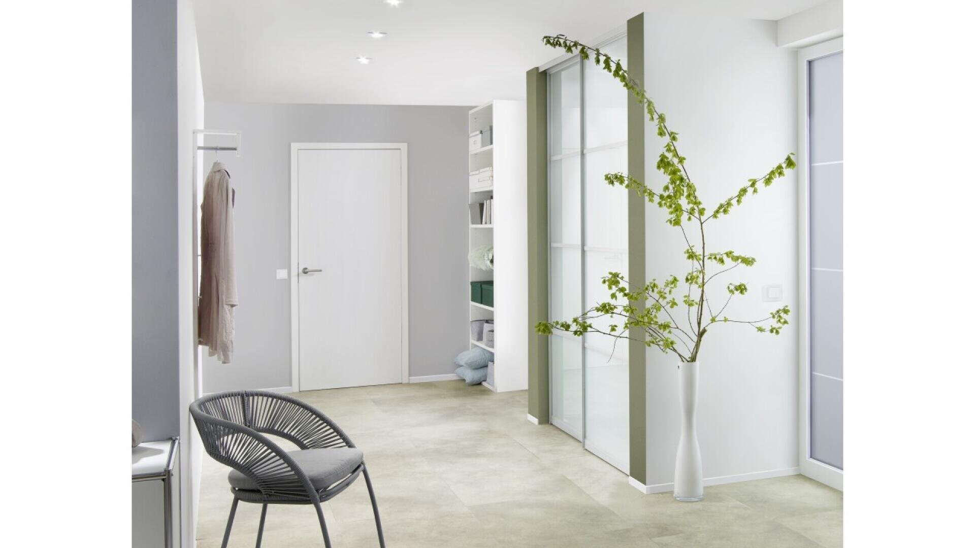 Couloir minimaliste avec des murs gris clair, une porte blanche fermée, une chaise grise moderne, un vase blanc contenant de hautes branches vertes et des portes coulissantes en verre dépoli. Des étagères avec des serviettes pliées sont visibles à l'arrière-plan.