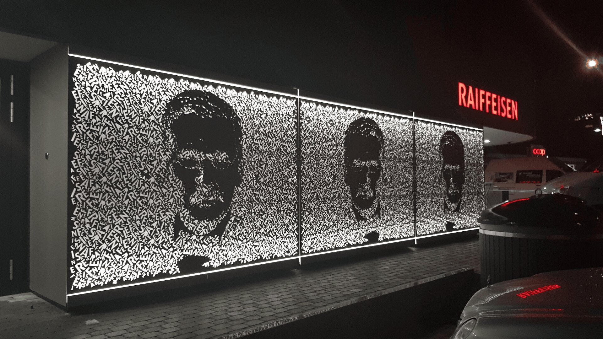Eine schwarz-weiß beleuchtete Wandmalerei bei Nacht zeigt drei stilisierte menschliche Gesichter, die sich aus abstrakten Formen zusammensetzen, und steht vor einer Raiffeisenbank. In der Nähe sind Autos und Straßenlaternen zu sehen.