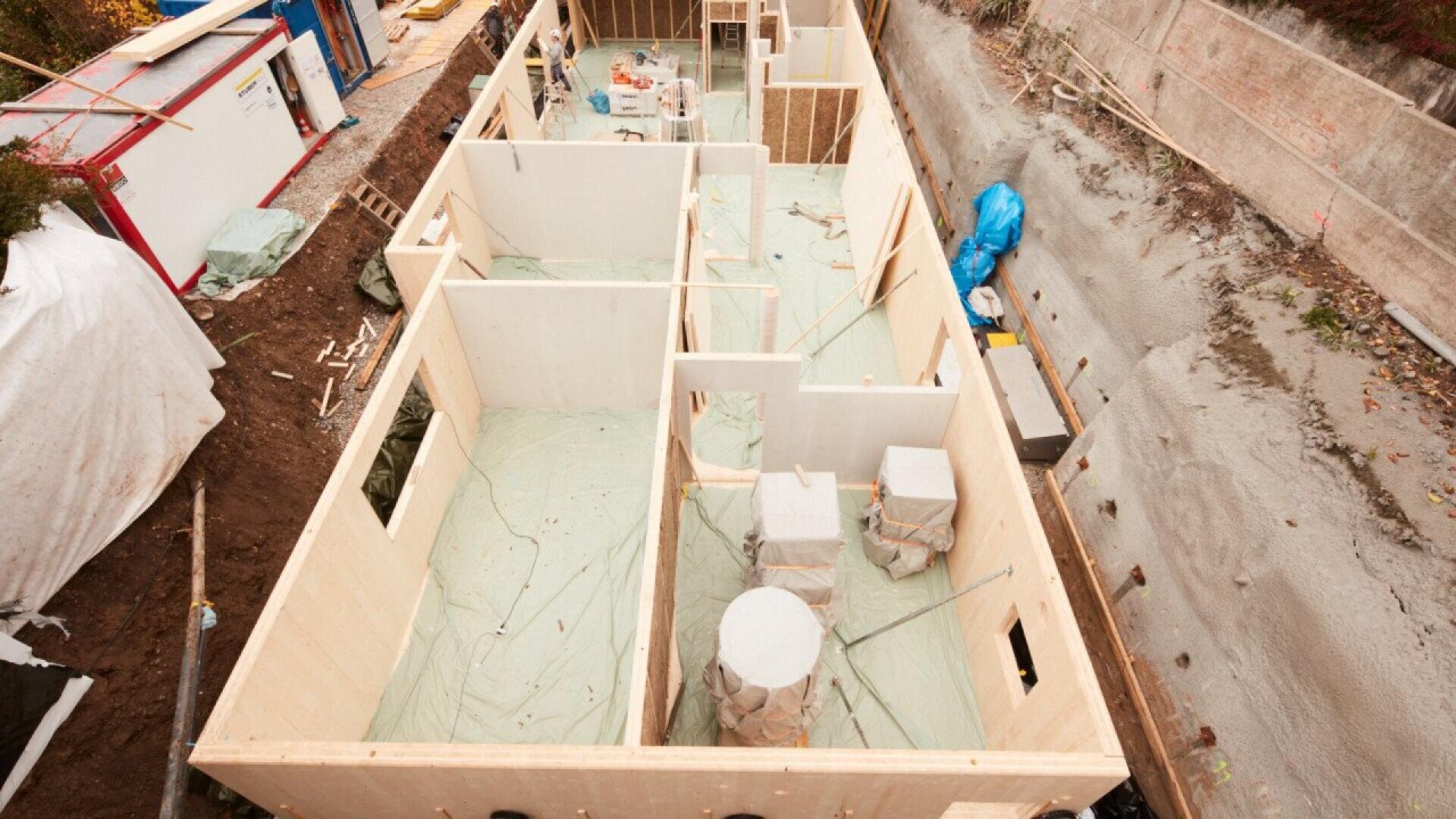Vue aérienne d'une maison en construction avec des murs en bois, des pièces partiellement achevées et des matériaux de construction éparpillés sur le site. Le sol environnant est irrégulier et couvert de bâches et d'outils de construction.