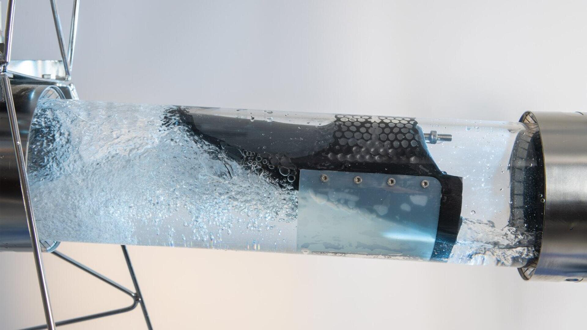 Un appareil sous-marin noir et bleu est présenté à l'intérieur d'un tube transparent rempli d'eau tourbillonnante, fixé à des supports métalliques de part et d'autre.