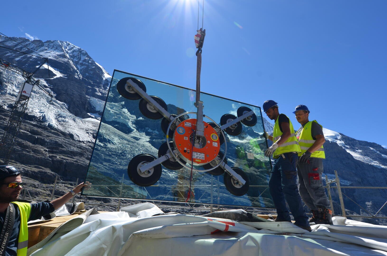 Zwei Arbeiter in Sicherheitswesten führen einen Kran, der eine große Glasplatte mit Saugnäpfen auf einer Baustelle anhebt, mit Bergen und einem klaren blauen Himmel im Hintergrund.