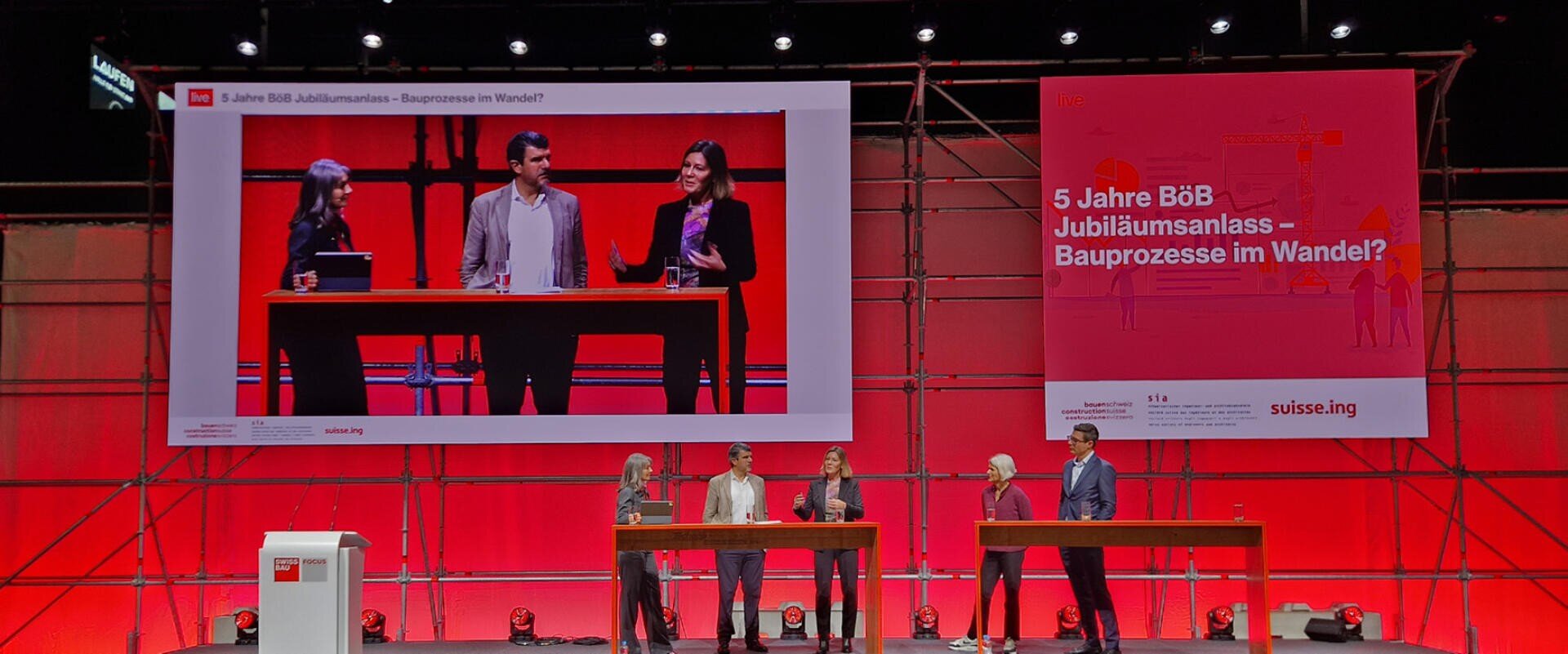 Fünf Personen stehen und sprechen auf der Bühne bei einer Podiumsdiskussion, über ihnen ist ein großer Bildschirm mit ihrem Bild zu sehen. Der Hintergrund und die Bühnenbeleuchtung sind rot, und auf einem Schild steht: "5 Jahre BÖB Jubiläumsanlass - Bauprozesse im Wandel".