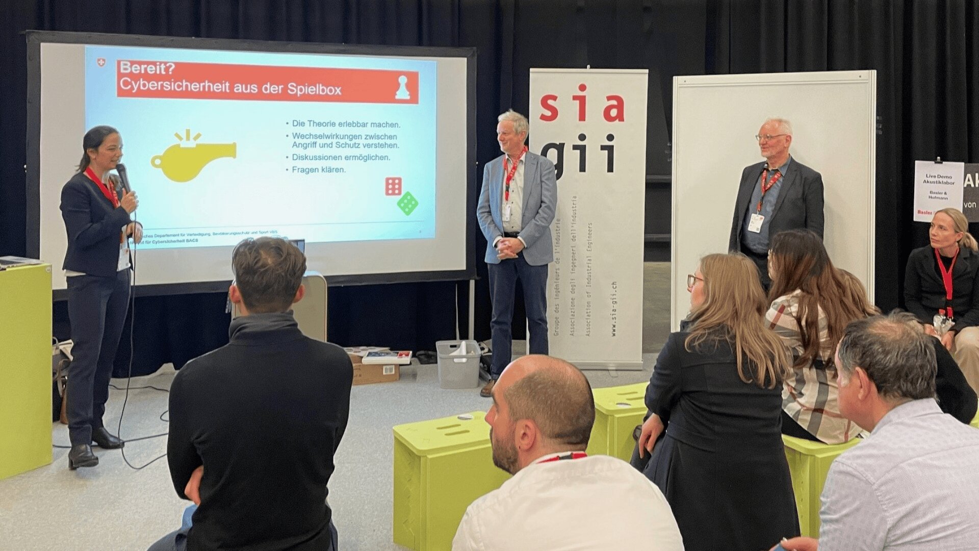 Eine Frau spricht in ein Mikrofon vor einer Präsentationsfolie über Cybersicherheit, während drei Personen auf der Bühne stehen und ein Publikum zuhört. Es scheint sich um eine Konferenz oder einen Workshop zu handeln, mit sichtbarer Beschilderung und Namensschildern.