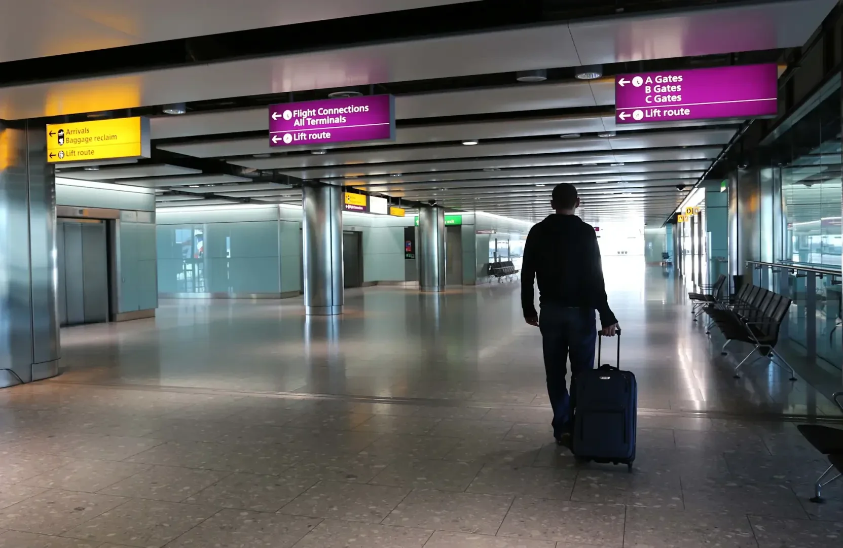 Eine Person geht durch ein leeres Flughafenterminal und zieht dabei einen Rollkoffer. Über den Köpfen zeigen Schilder den Weg zu Ankünften, Flügen, Gates und Aufzügen. Sitze und Glaswände säumen das Terminal.