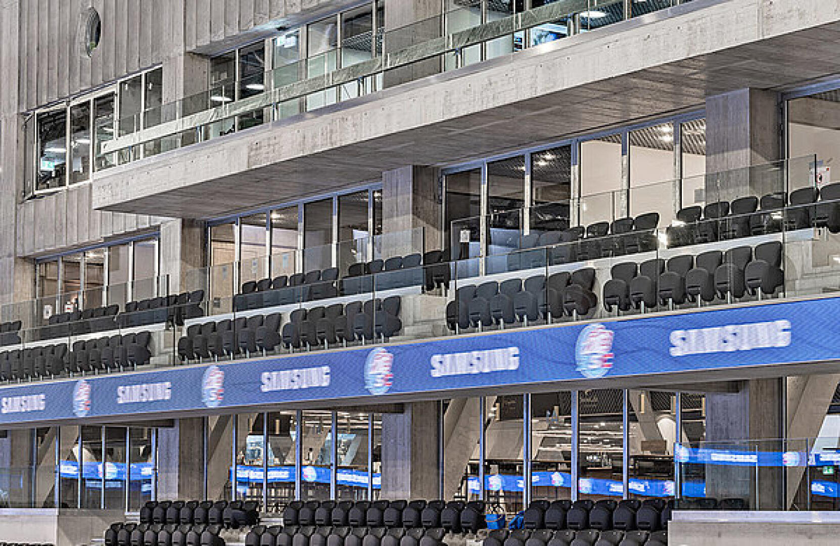 Des rangées de sièges noirs vides, entourés de parois vitrées, surplombent une salle omnisports. Une bannière numérique bleue, le long de la façade, affiche le logo Samsung à plusieurs reprises. La structure en béton domine l'arrière-plan.