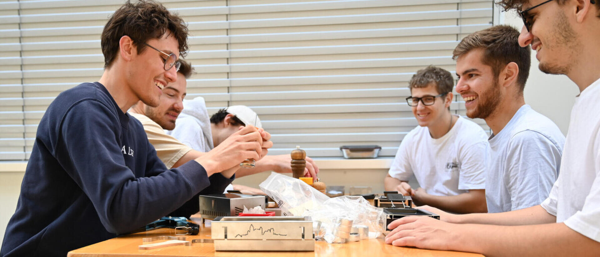 Fünf junge Männer sitzen lächelnd um einen Tisch und arbeiten gemeinsam an einem Projekt mit Werkzeugen und kleinen mechanischen Teilen; die Atmosphäre wirkt kooperativ und freundlich.