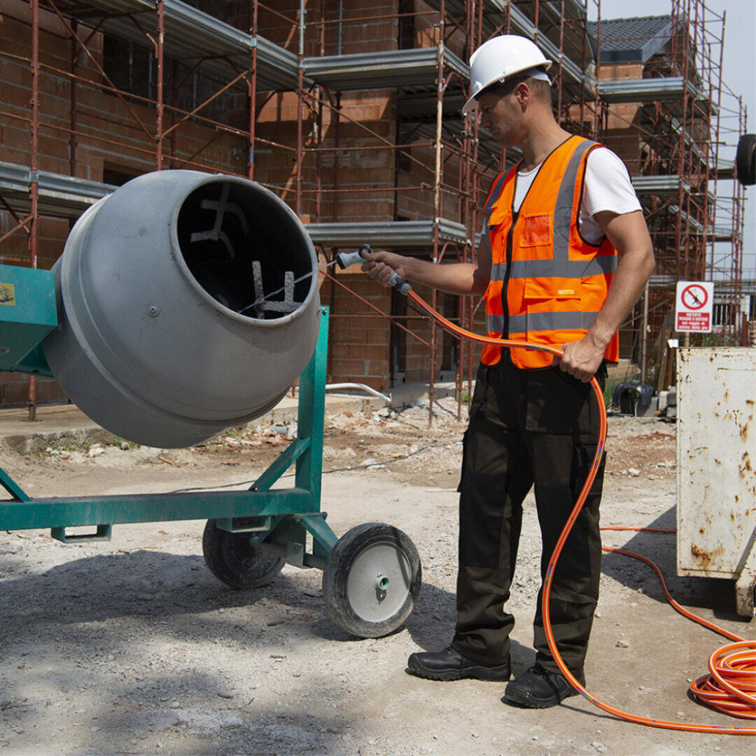 Ein Bauarbeiter mit weißem Schutzhelm und orangefarbener Sicherheitsweste reinigt mit einem Schlauch einen Zementmischer auf einer Baustelle mit einem Baugerüst im Hintergrund.