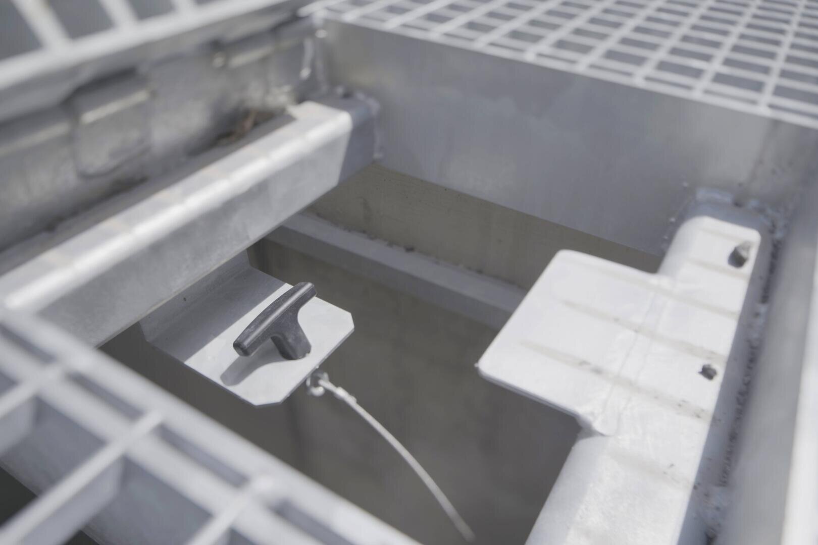 Nahaufnahme einer industriellen Metallstruktur mit Gitterboden, einer rechteckigen Öffnung und einem T-förmigen Griff, der an einem Kabel innerhalb der Öffnung befestigt ist. Die Gesamtfarbe ist metallisch grau.