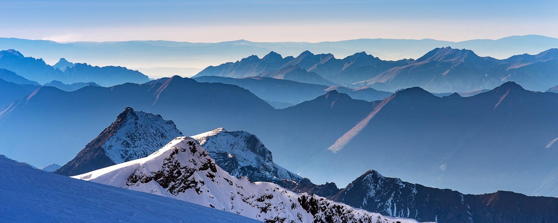 Schneebedeckte Berggipfel im Vordergrund mit überlagerten blauen Graten und weit entfernten Bergen, die in einem nebligen Himmel verblassen.