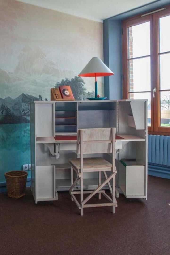 Un petit bureau en bois avec des étagères ouvertes et des livres, une lampe rouge et blanche et une chaise en bois se trouvent devant une fenêtre éclairée par la lumière naturelle, à côté d'une poubelle et d'un radiateur. Une peinture murale orne le mur derrière le bureau.