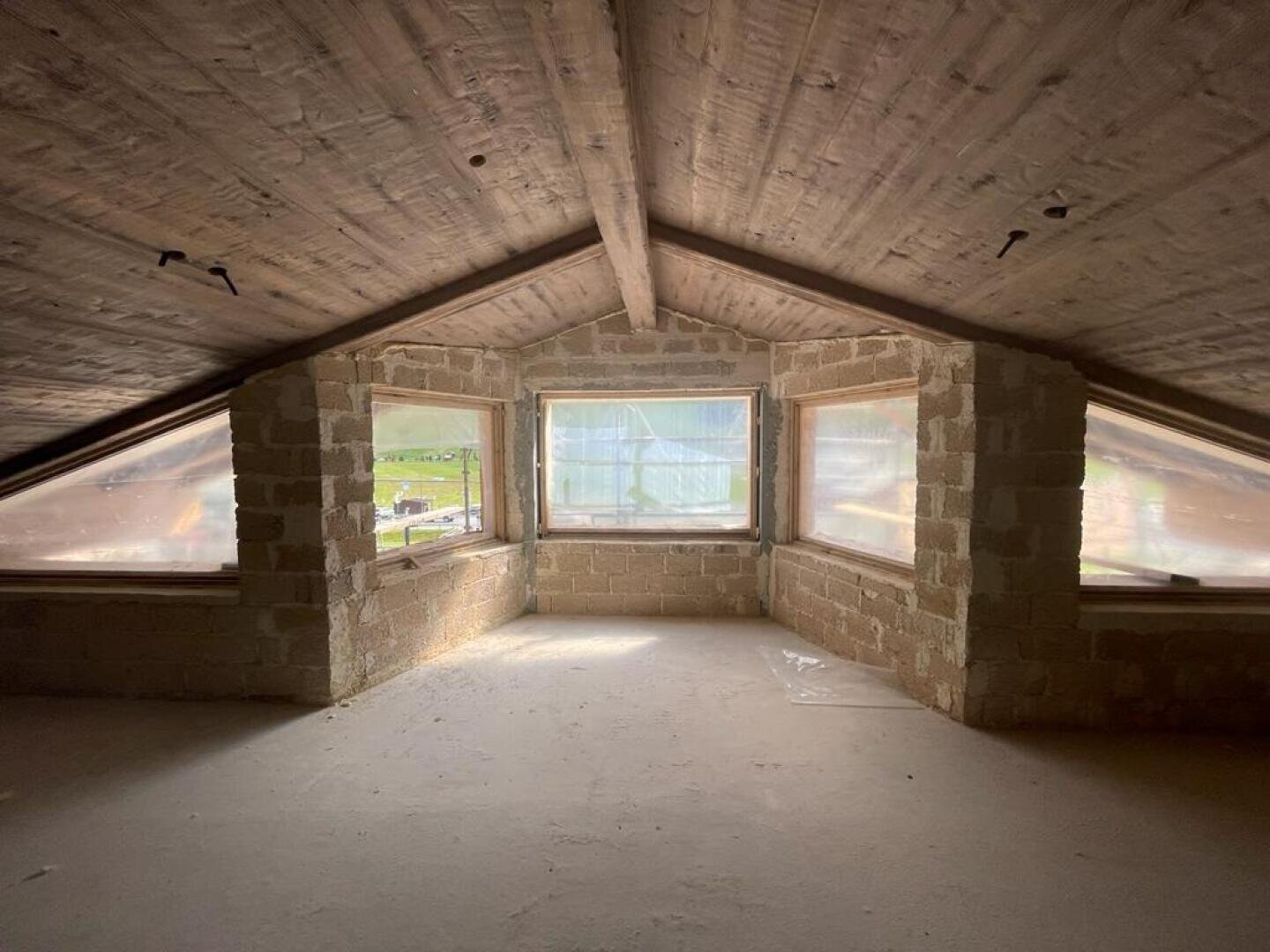 Ein geräumiger Dachboden mit unfertigen Ziegelwänden, Holzbalken an der Decke und drei großen Fenstern, die viel natürliches Licht hereinlassen. Der Boden ist staubig und die Fenster bieten einen Blick auf das Grün draußen.