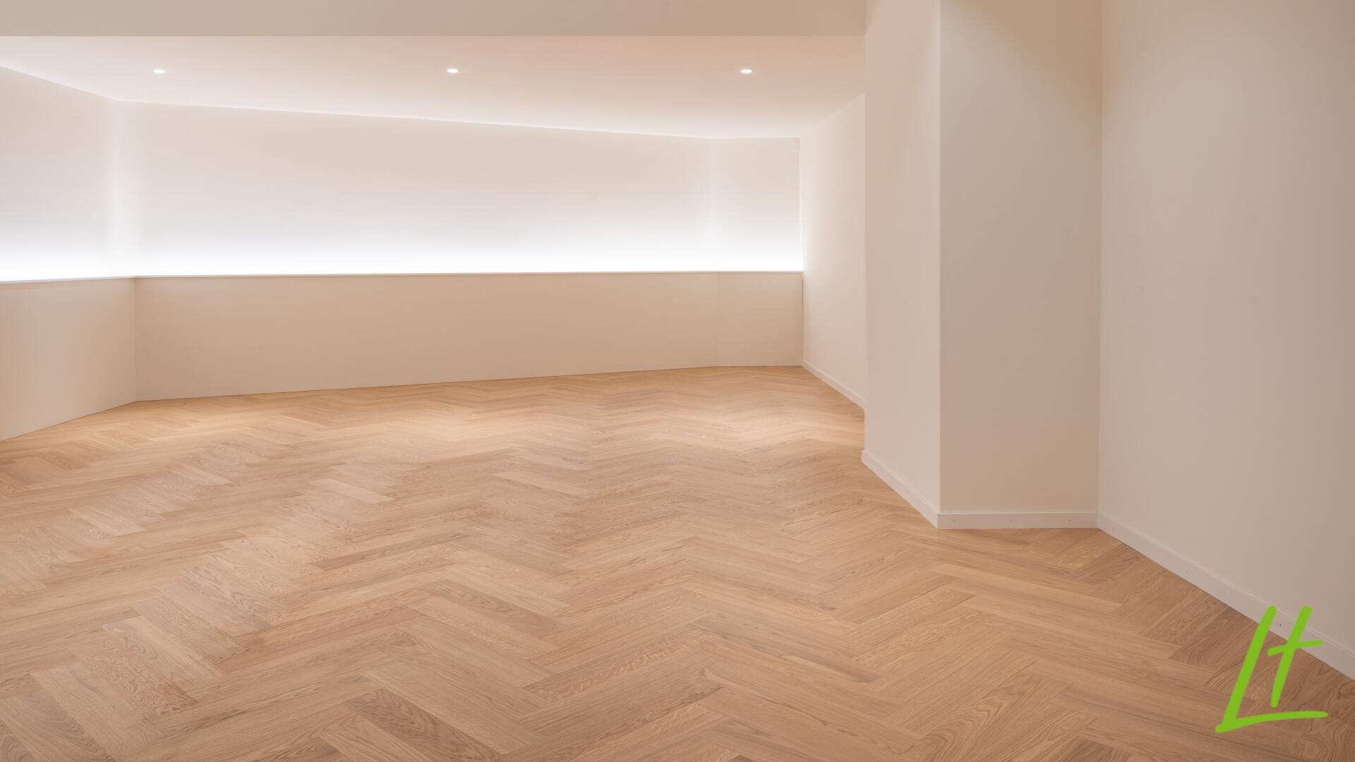 Minimalistischer, leerer Raum mit hellem Holzboden im Fischgrätmuster, weißen Wänden, Deckeneinbauleuchten und einem Streifen mit sanfter Beleuchtung an der hinteren Wand. Das grüne Logo befindet sich in der unteren rechten Ecke.
