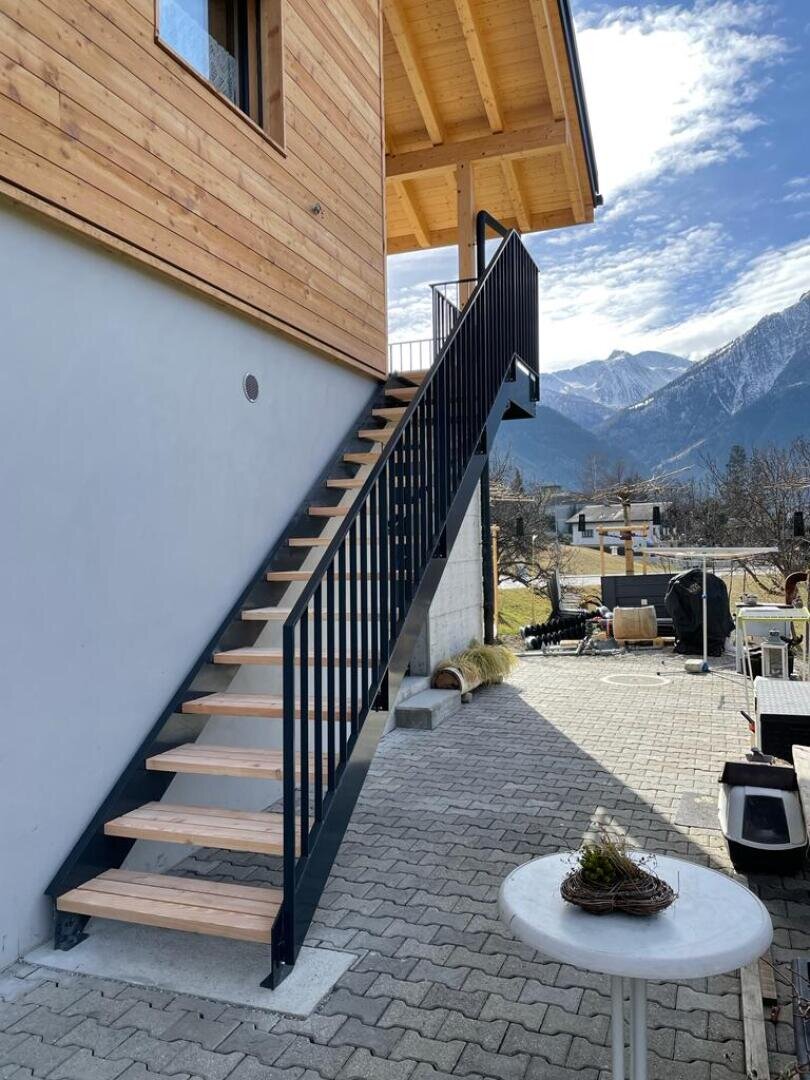 Außentreppe aus Holz und Metall, die an ein modernes Haus angebaut ist und in ein Obergeschoss führt. Die Treppe blickt auf eine gepflasterte Terrasse mit einem runden Tisch und einem malerischen Blick auf die Berge unter einem teilweise bewölkten Himmel.
