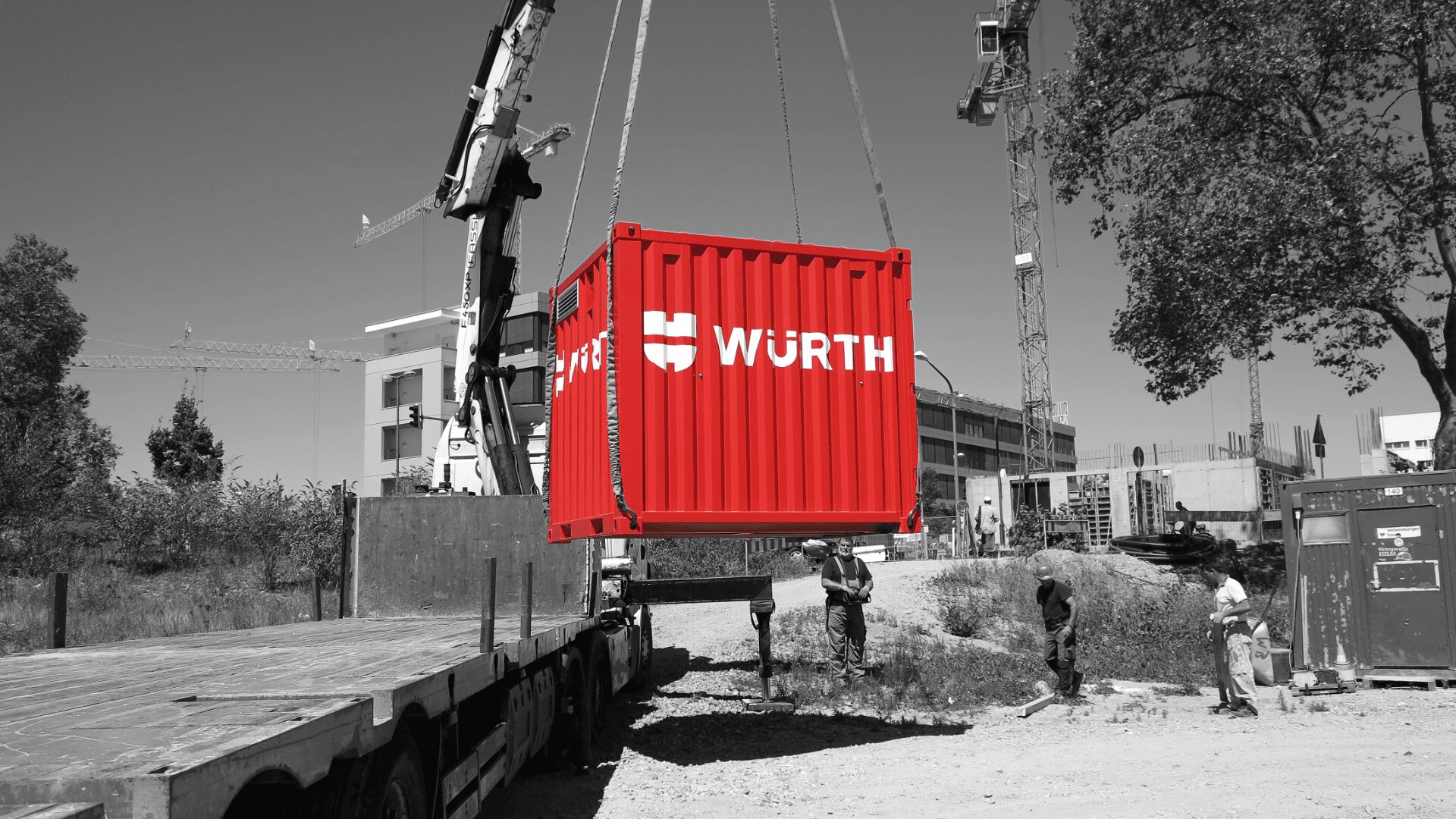 Ein leuchtend roter Schiffscontainer mit dem WÜRTH-Logo wird auf einer Baustelle von einem Kran angehoben, während Arbeiter in der Nähe sind. Der rote Container hebt sich von dem schwarz-weißen Hintergrund ab.
