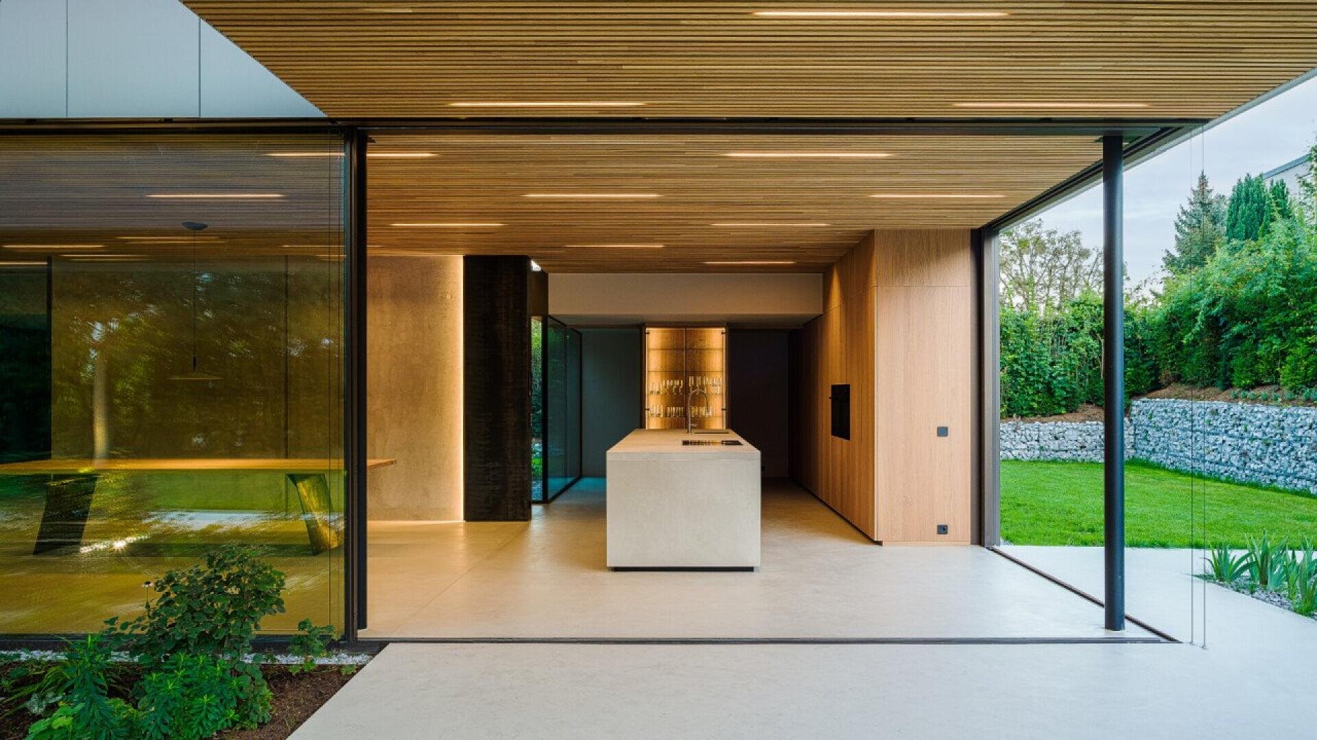 Cuisine moderne avec îlot en béton, murs en bois et portes vitrées ouvrant sur une pelouse et un jardin verdoyants.