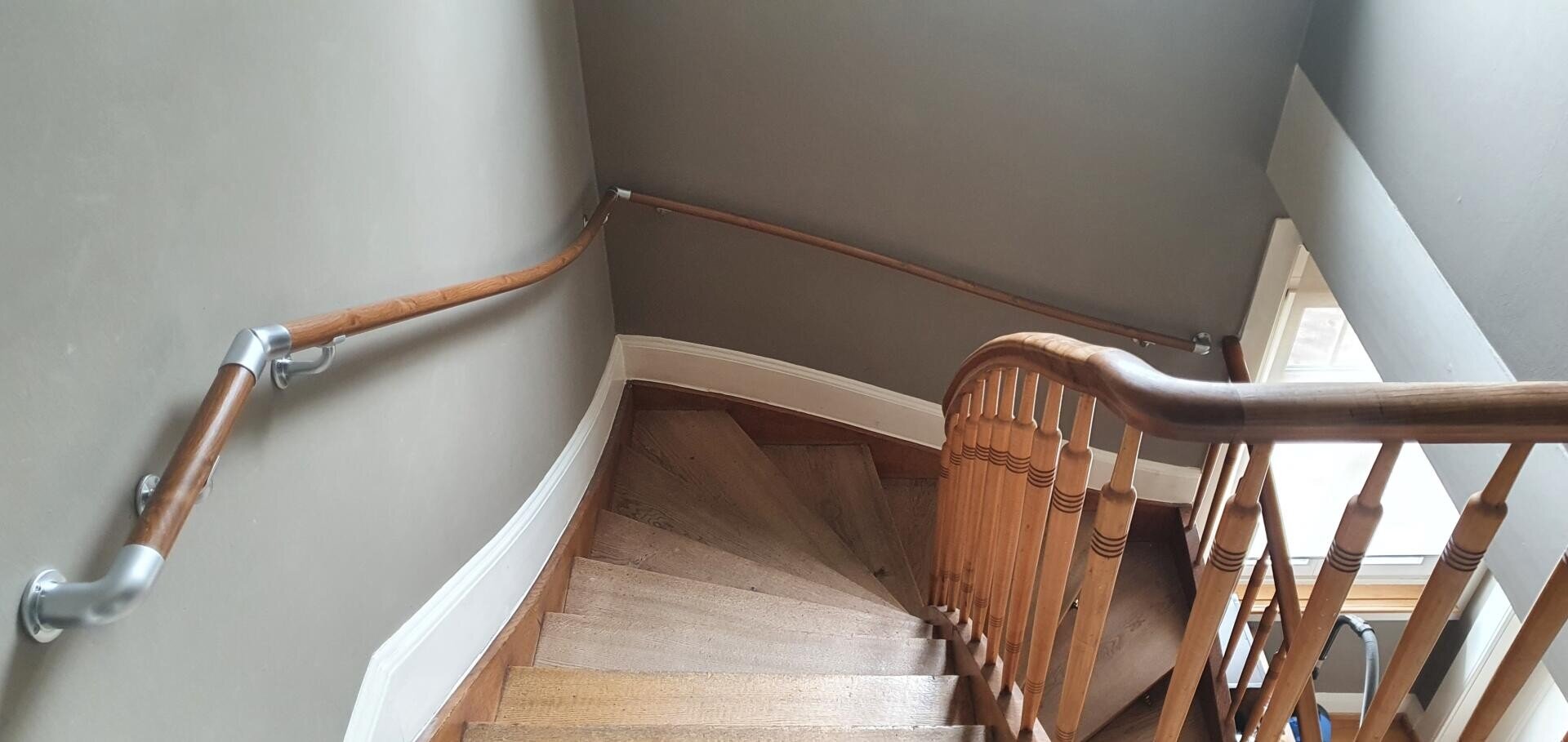 Eine Holztreppe mit geschwungenem Geländer führt entlang einer grauen Wand mit hölzernem Handlauf hinunter in ein unteres Stockwerk. Durch ein Fenster am unteren Ende der Treppe fällt natürliches Licht ein.