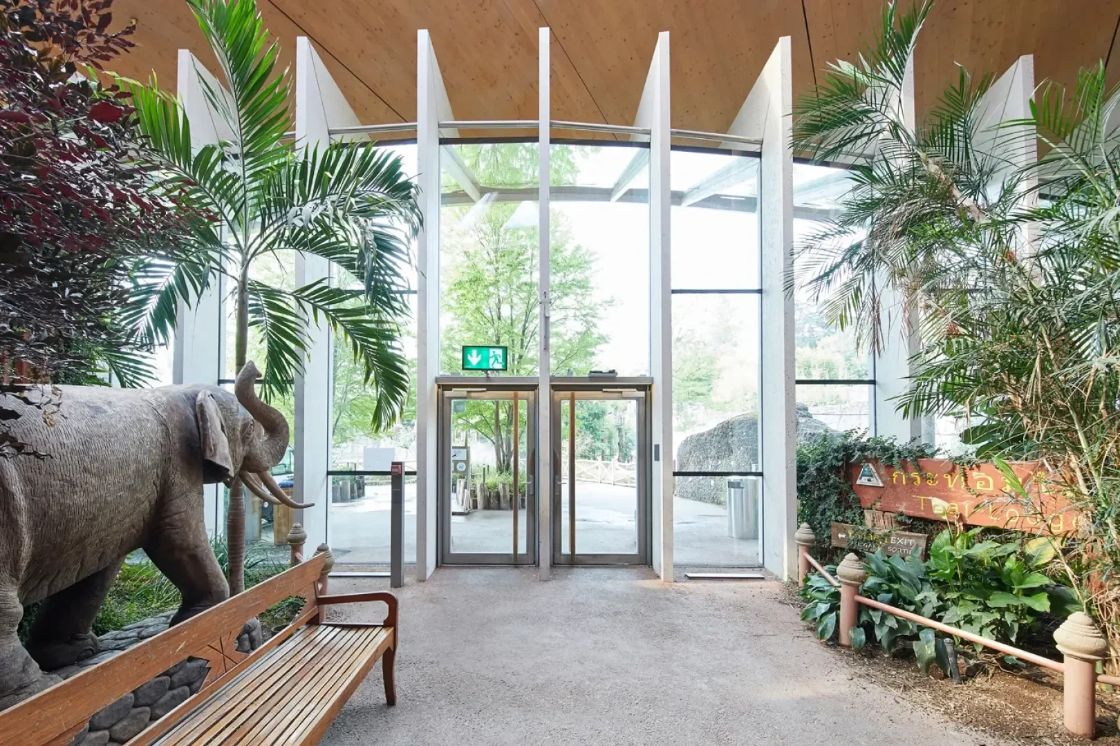 Geräumiger Innenbereich mit großen Glastüren und hohen Fenstern, flankiert von tropischen Pflanzen und einer Elefantenstatue. Links steht eine Holzbank, rechts ein Holzschild mit thailändischer Schrift.
