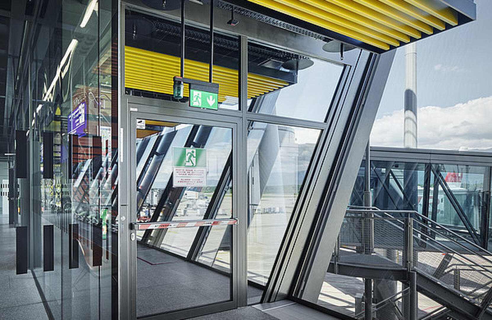 Modernes Flughafenterminal mit großen Glasfenstern, einer Glastür mit einem grünen Ausgangsschild und gelben Deckenplatten. Durch das Glas sind Spiegelungen und Außenansichten der Landebahn und des Himmels zu sehen.