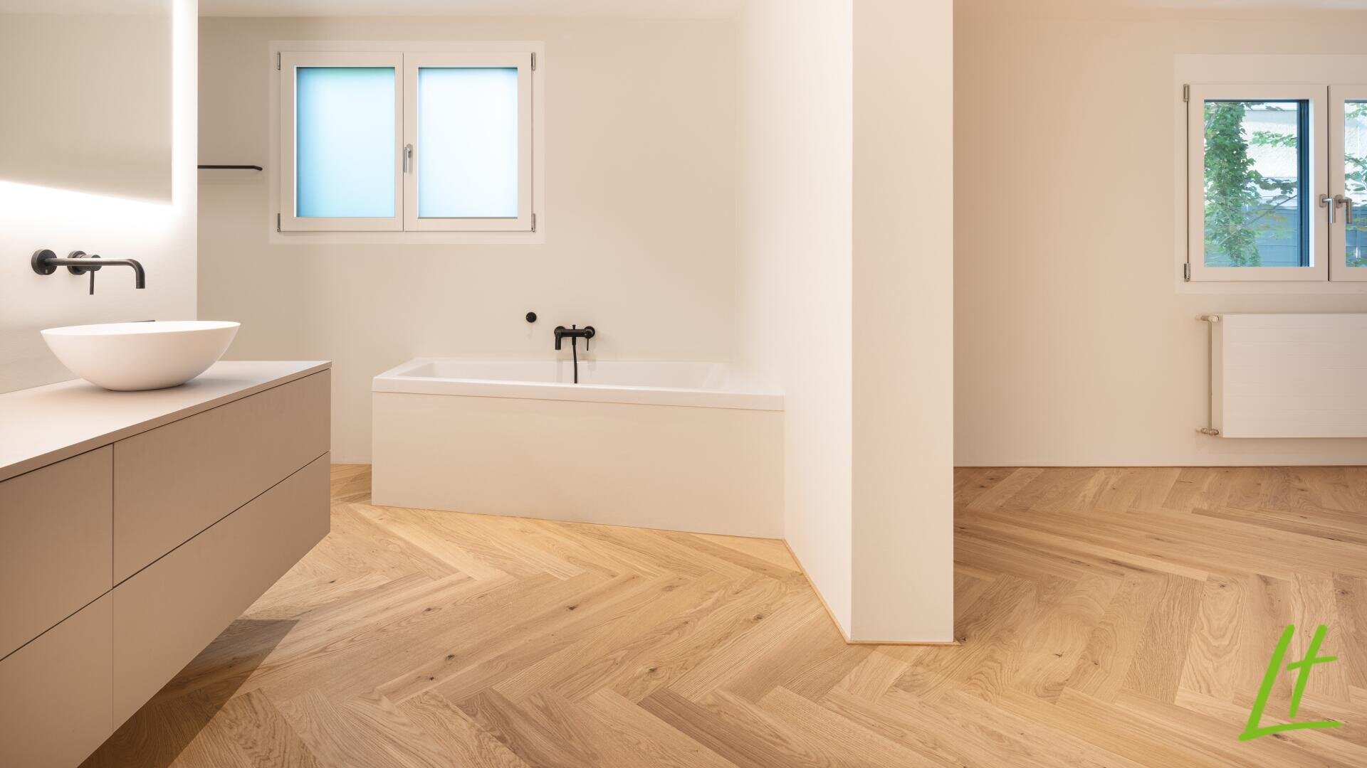 Salle de bains minimaliste avec un sol en bois clair à chevrons, une baignoire autoportante avec des accessoires noirs, un meuble-lavabo flottant avec un évier en forme de récipient, deux fenêtres givrées et des murs blancs.