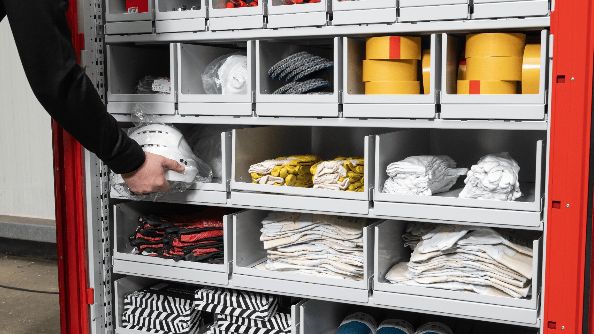 Die Hand einer Person holt einen weißen Schutzhelm aus einem Regal in einem Schrank, der mit ordentlich geordneter Sicherheitsausrüstung wie Handschuhen, gerolltem gelbem Klebeband, gerollten Handtüchern und gefalteten Uniformen gefüllt ist.