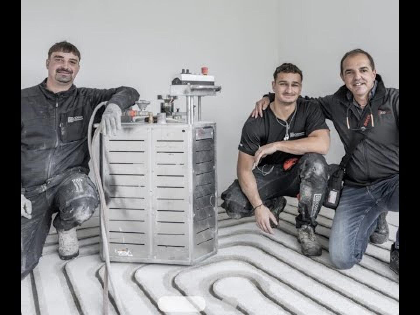 Trois hommes en vêtements de travail sont agenouillés sur un sol avec des tuyaux de chauffage par le sol exposés et sourient à la caméra. Ils posent à côté d'une machine métallique utilisée pour l'installation ou le contrôle du système de chauffage.
