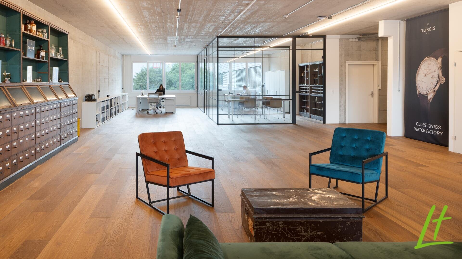 Bureau moderne en open space avec plancher en bois, plafond en béton apparent, fauteuils lounge, canapé vert, armoires anciennes et salle de réunion vitrée. Deux personnes travaillent à des bureaux situés près de grandes fenêtres en arrière-plan.